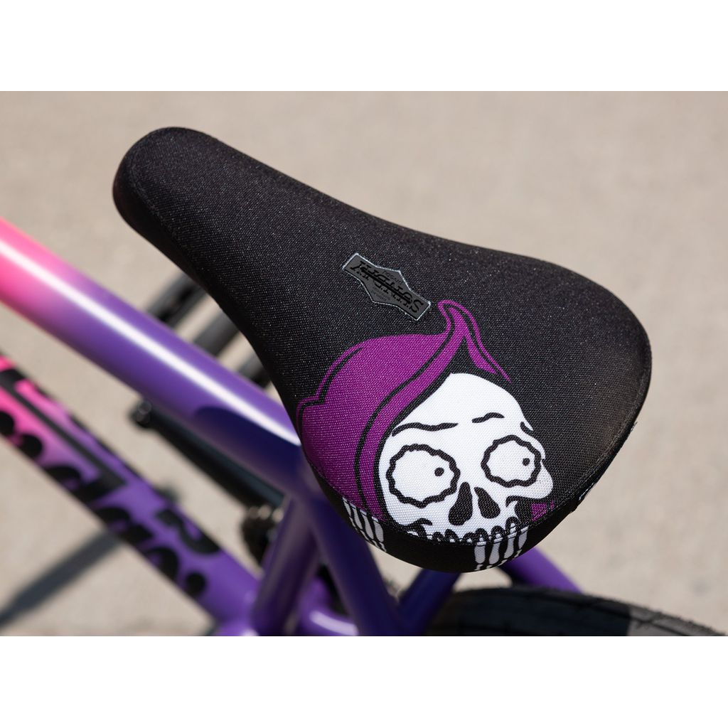 Close-up of a BMX bike seat featuring a skull graphic with a purple hood. The seat is mounted on the striking chromoly frame of the Sunday Street Sweeper 20 Inch Bike, which is painted in vibrant purple and pink hues.