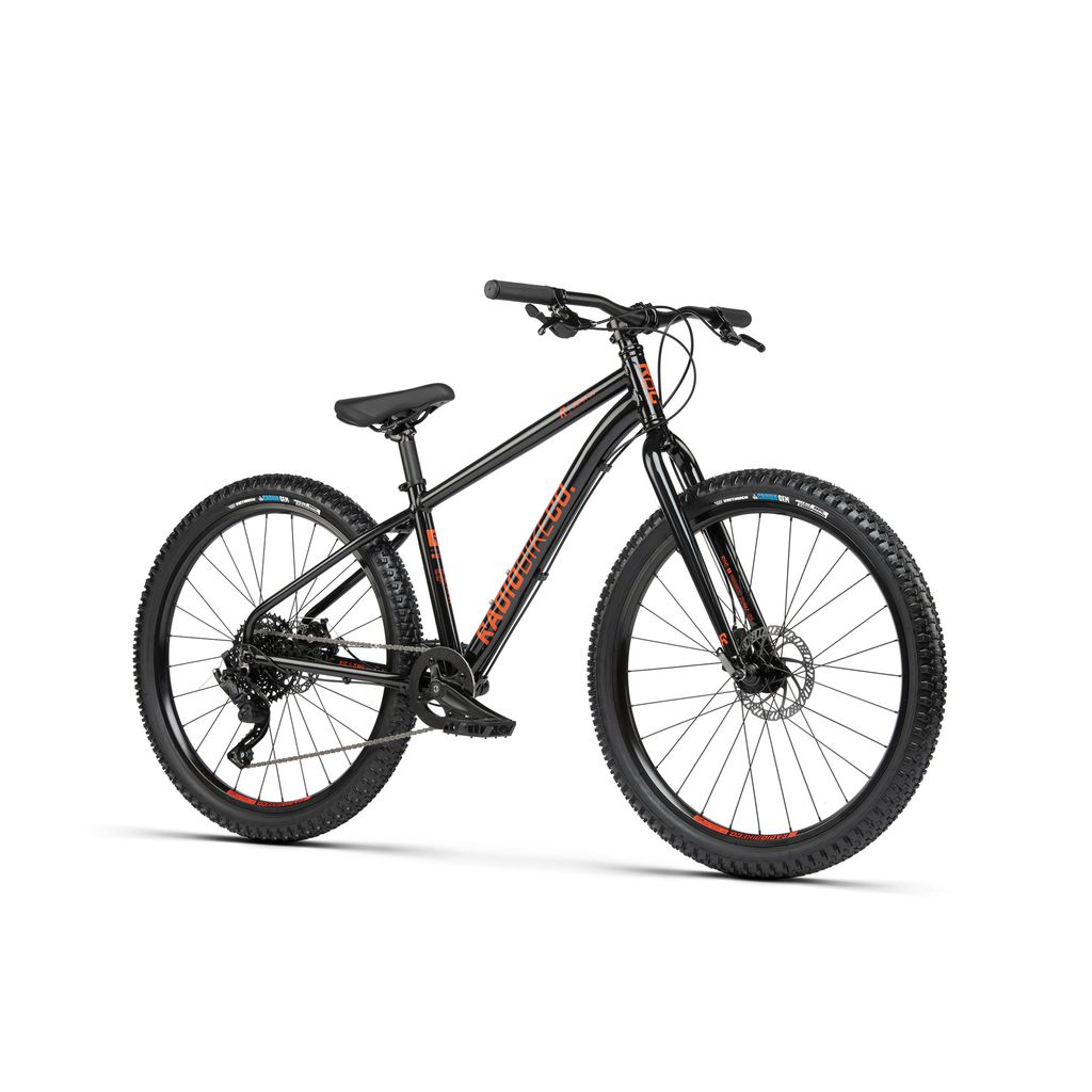 The Radio 26 Inch Zuma Bike is a black mountain bike with a lightweight alloy frame featuring orange accents. It includes front suspension, thick tires, and disc brakes. The bike is equipped with a flat handlebar and a padded seat, making it an ideal Kids MTB bike designed with child-specific components.