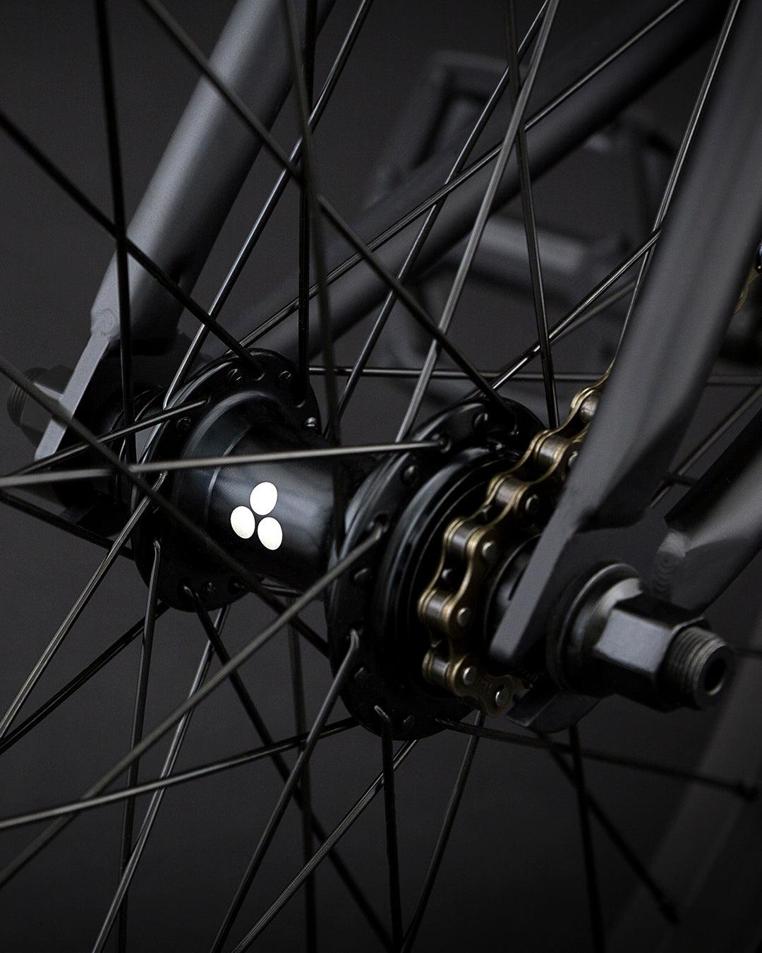 A close-up of the Fly Bikes Neutron 20 Inch Bike's rear wheel hub features a logo, highlighting its dark spokes and chainset against a dark background. The CrMo frame enhances durability and strength, contributing to the bike's sleek design.