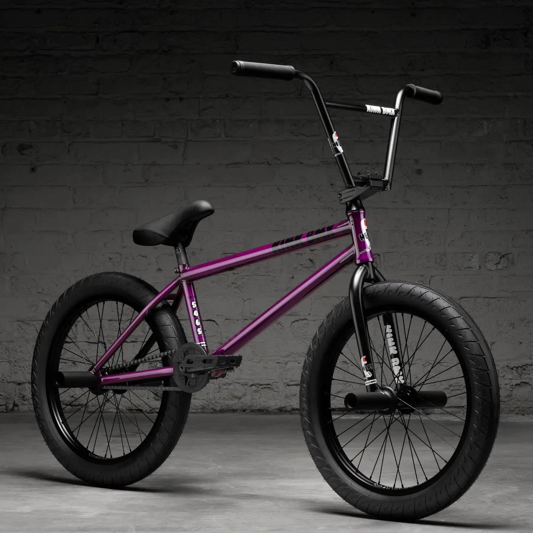 The Kink Williams 20 Inch Bike (2026), featuring a purple frame, black handlebars, and black tires, is set against a gray brick wall backdrop.