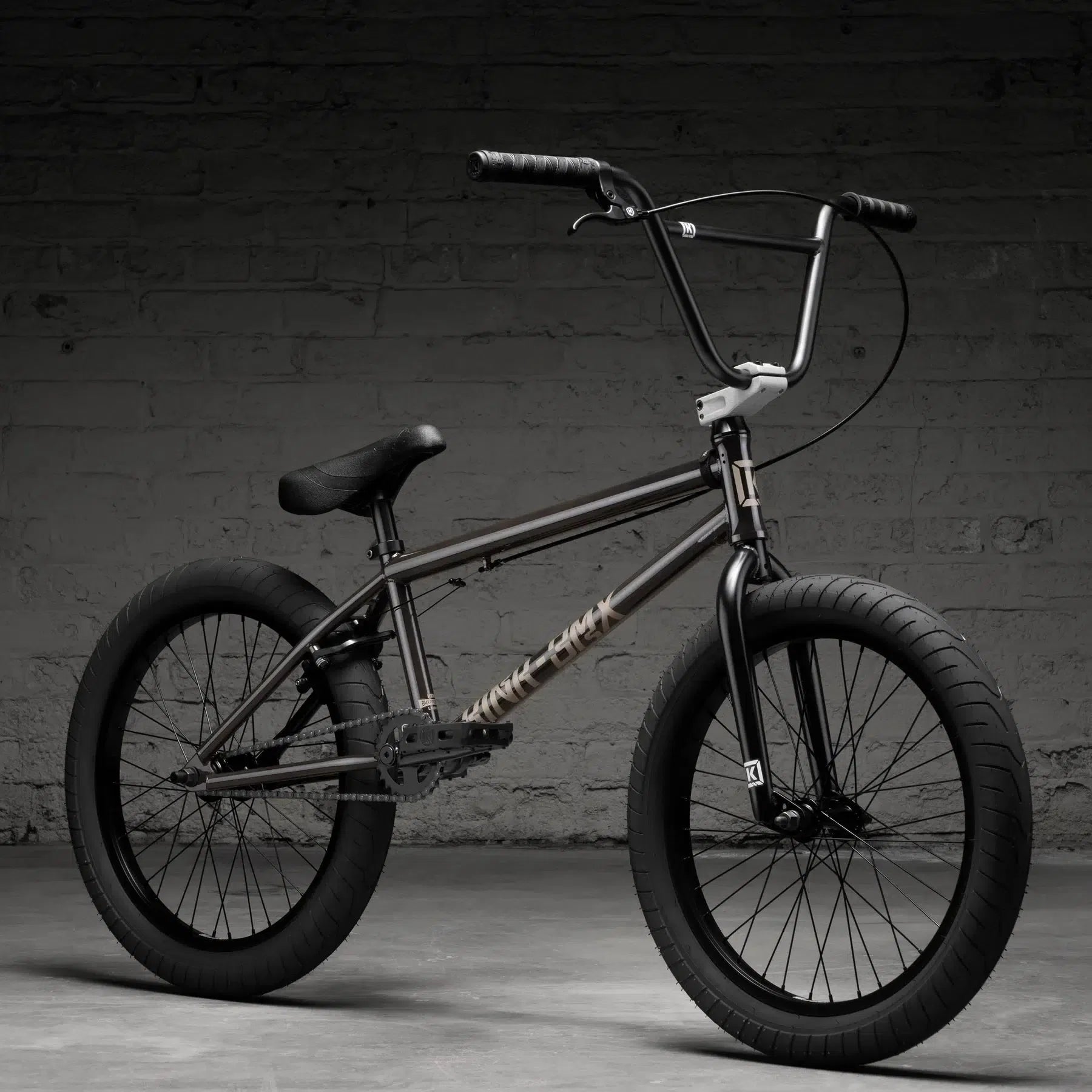 A Kink Launch 20 Inch BMX bike from 2026 featuring wide tires, a graffiti-style logo, sealed bearings, and durable Mission parts leans against a dark brick wall for smooth rides.