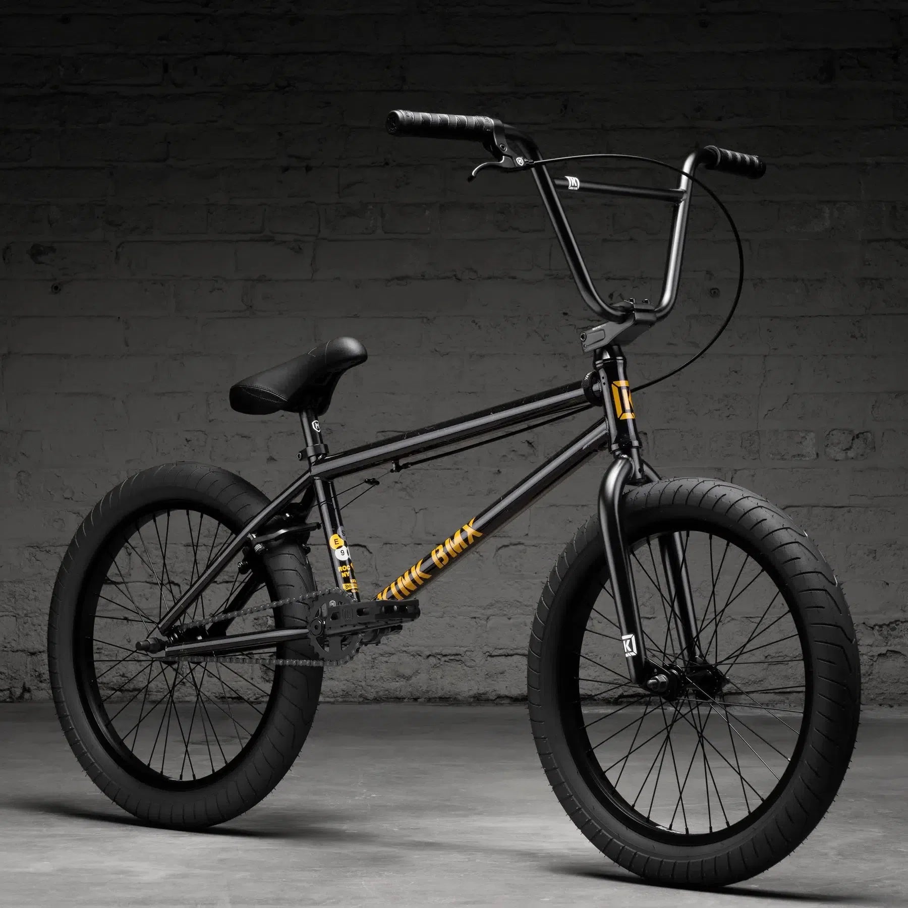 The Kink Gap XL 20 Inch Bike (2026) in black, featuring thick tires and a gold logo, rests against a gray brick wall on the concrete floor.