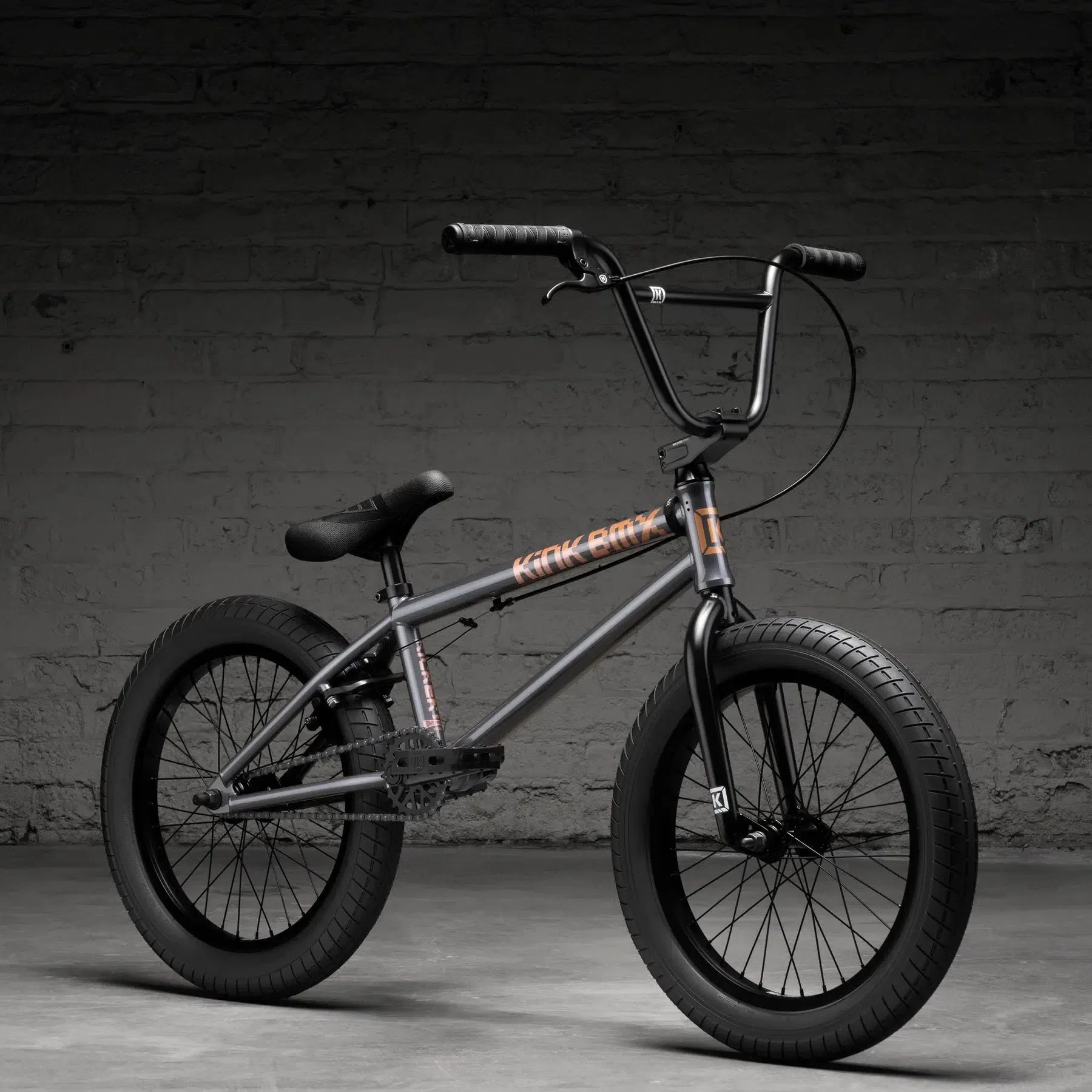 A true Kink Kicker 18 Inch Bike (2026) with a matte black frame, thick tires, and high handlebars stands against a gray brick wall on a concrete floor.