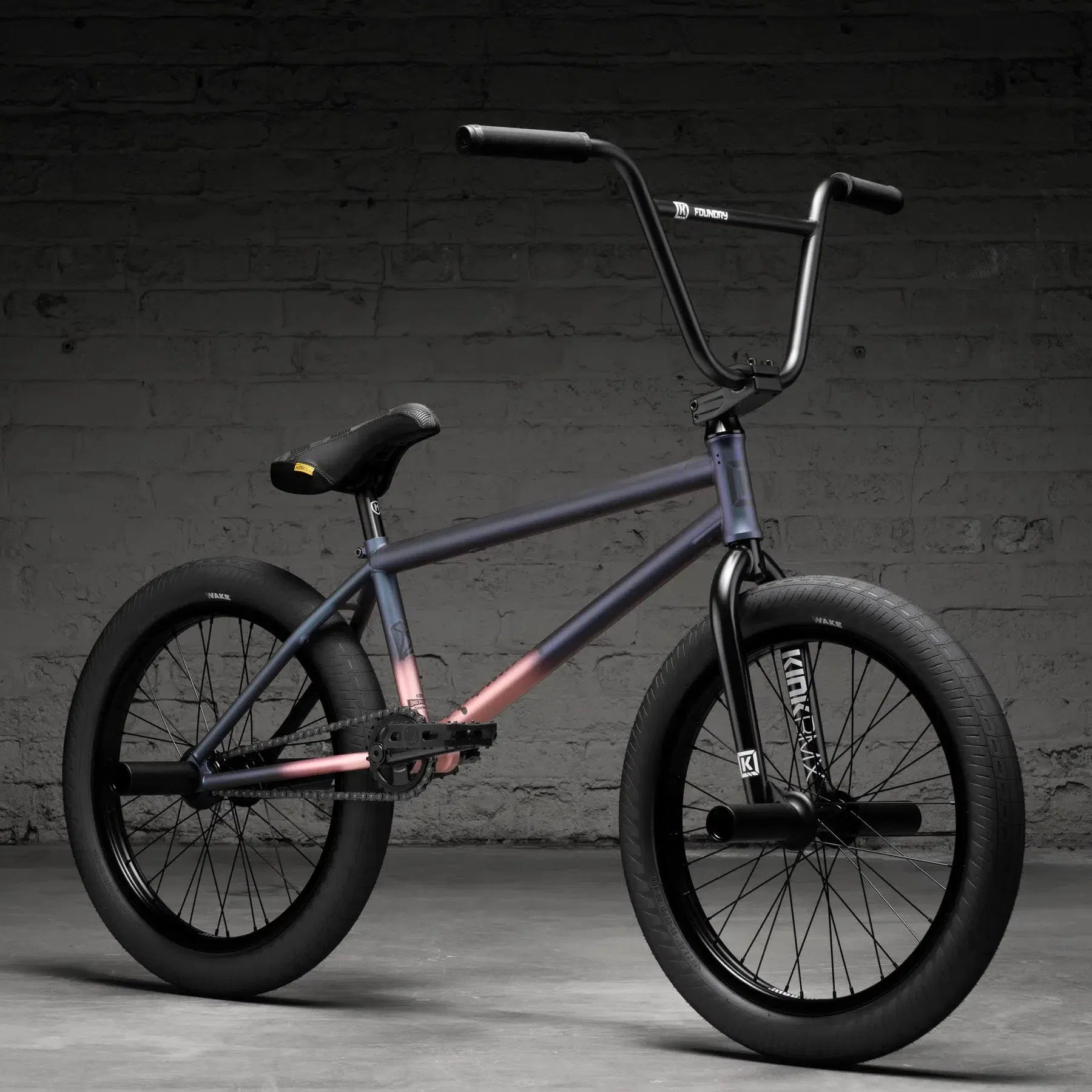 The Kink Downside 20 Inch Bike (2026), featuring a gradient frame, black tires, and high handlebars, is set against a concrete wall backdrop.