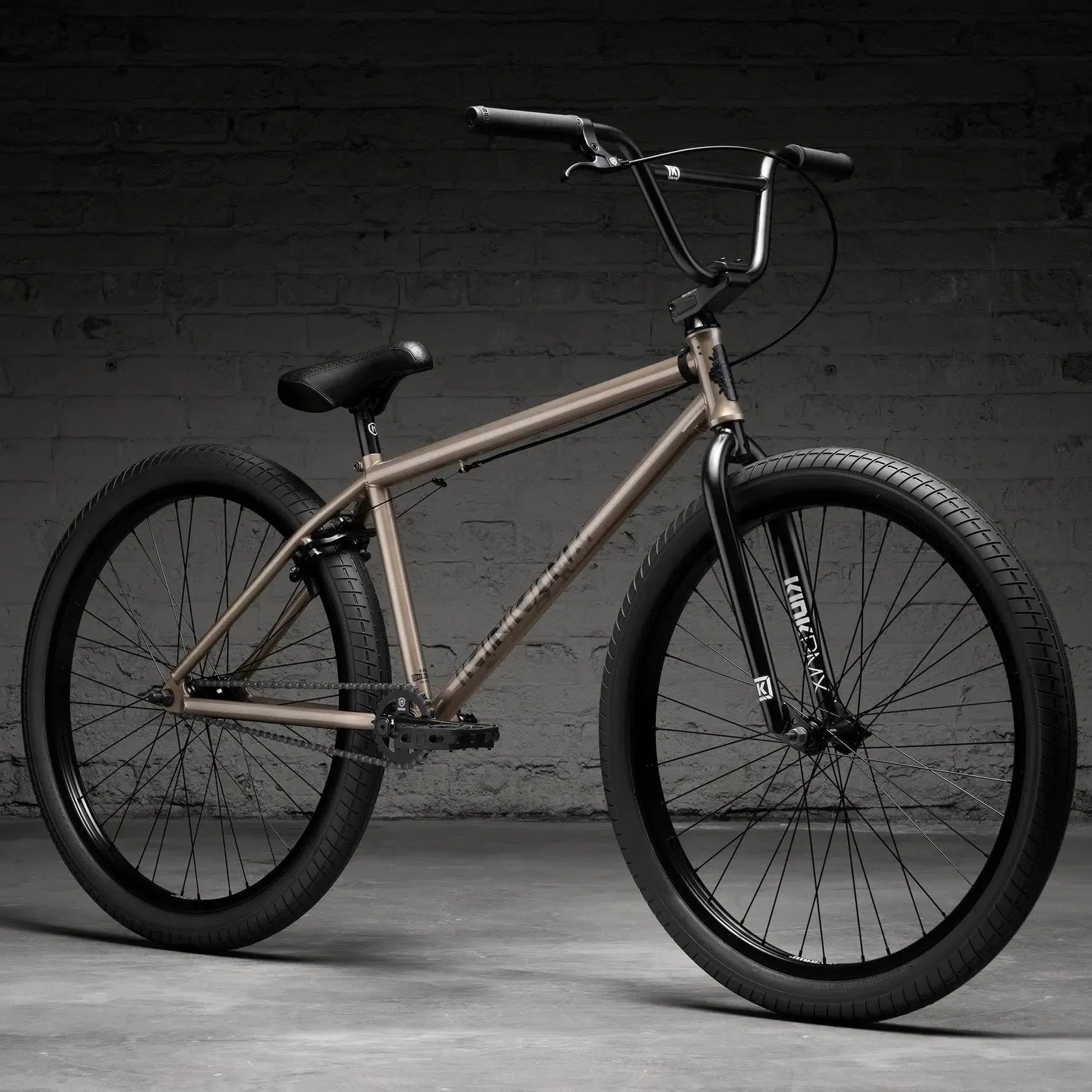 The Kink Drifter 26 Inch Bike (2026), featuring a beige BMX-style and robust Chromoly frame with large black tires, is positioned against a gray brick wall.