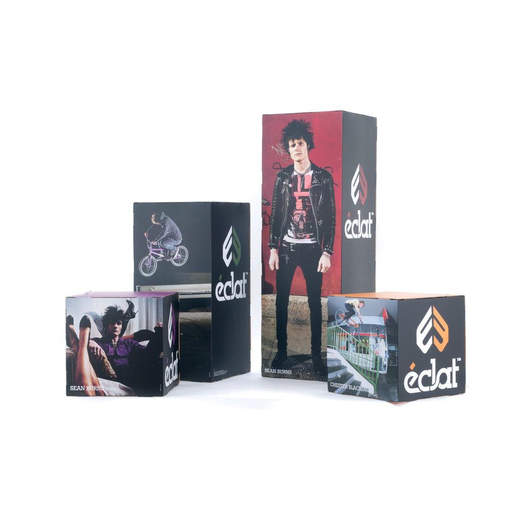 The Eclat Promo Stand Setup features four distinct éclat product boxes, each displaying images of a person standing, cycling, and sitting with the éclat logo prominently visible. Draw inspiration from team riders like Sean Burns through this exclusive presentation.