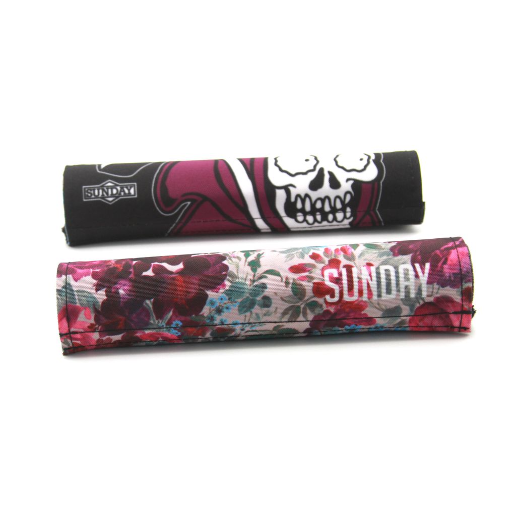 Two cylindrical fabric items with the brand name "Sunday." One of them, the Sunday Wallflower V2 Reversible Handlebar Pad, is reversible with a skull and dark design on one side and a floral pattern on the other, featuring a convenient Velcro closure for easy adjustments.