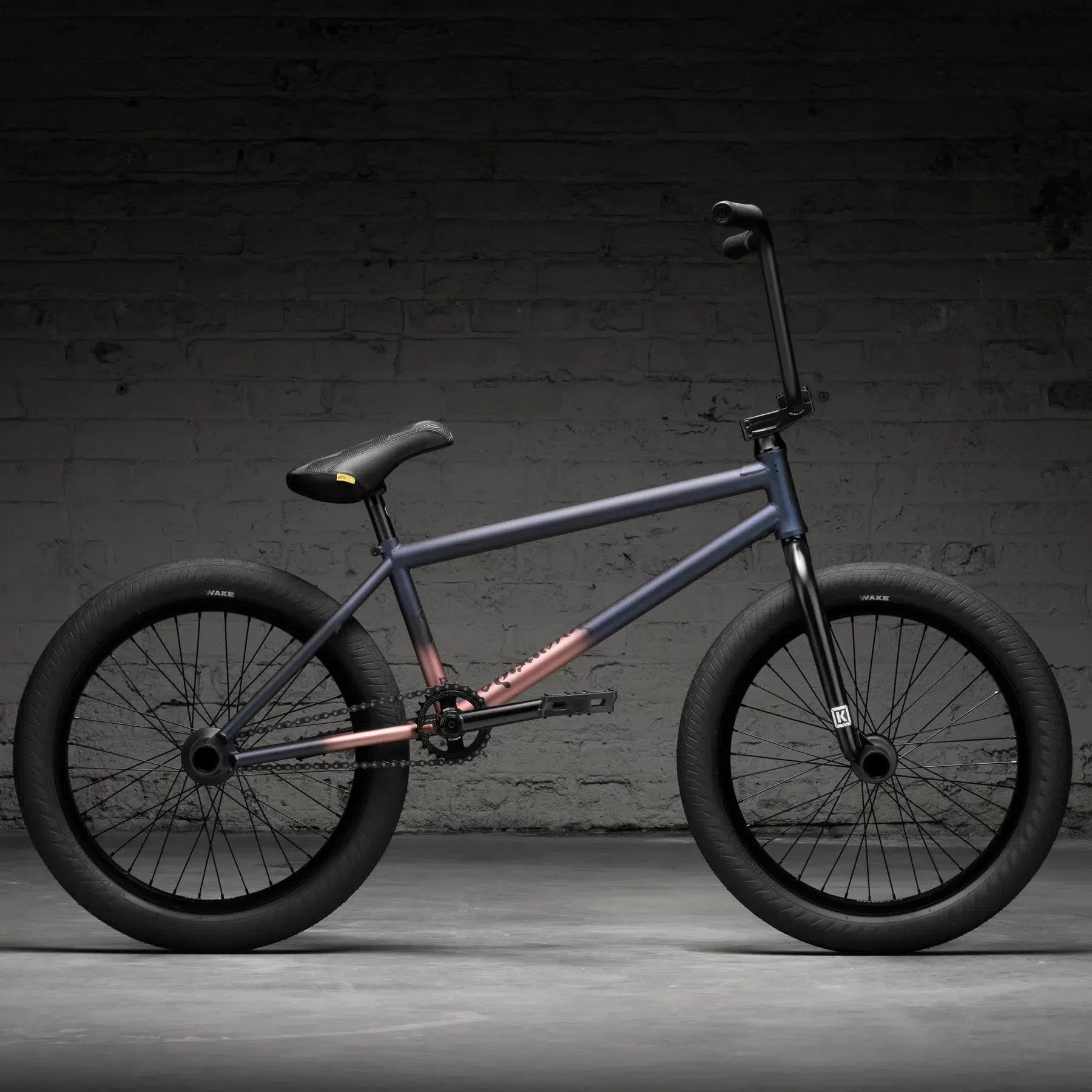 The Kink Downside 20 Inch Bike (2026) stands against a dark brick wall with its matte grey and pink frame. It features durable chromoly construction, black tires, a padded seat, and a straight handlebar, making this BMX truly exceptional.