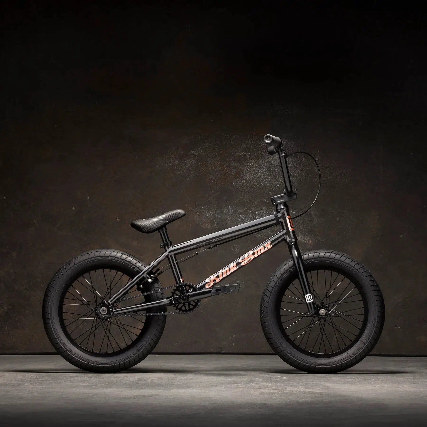 The Kink Carve 16 Inch Bike, with its black BMX style, highlights its simple frame and small seat with thick tires and Mission parts against a dark background.