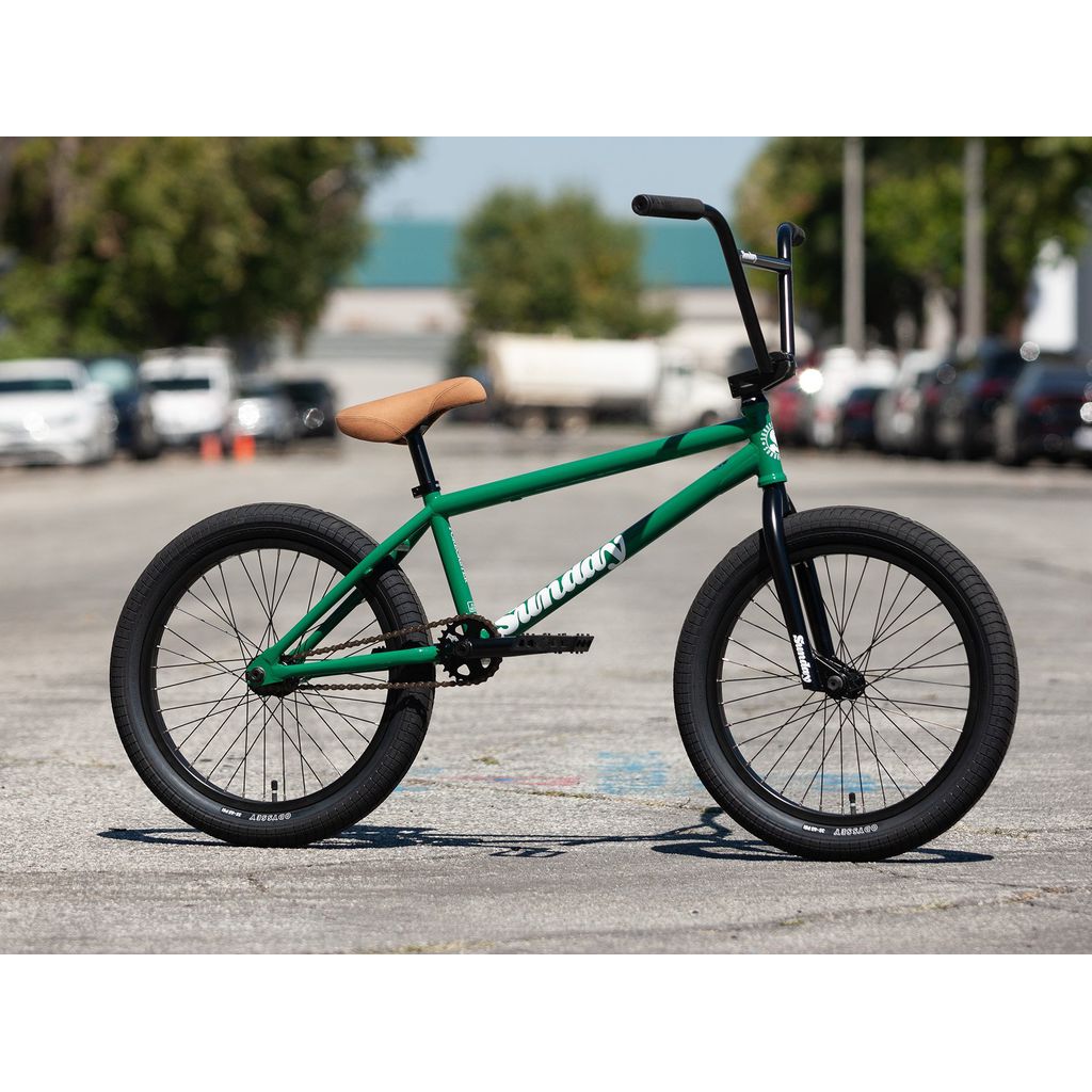 A Sunday Forecaster 20 Inch Bike (2022) / Alec Siemon model, featuring a green BMX frame with black tires and a brown seat, made with a 100% chromoly frame, is parked on a paved outdoor area with trees and vehicles in the background.