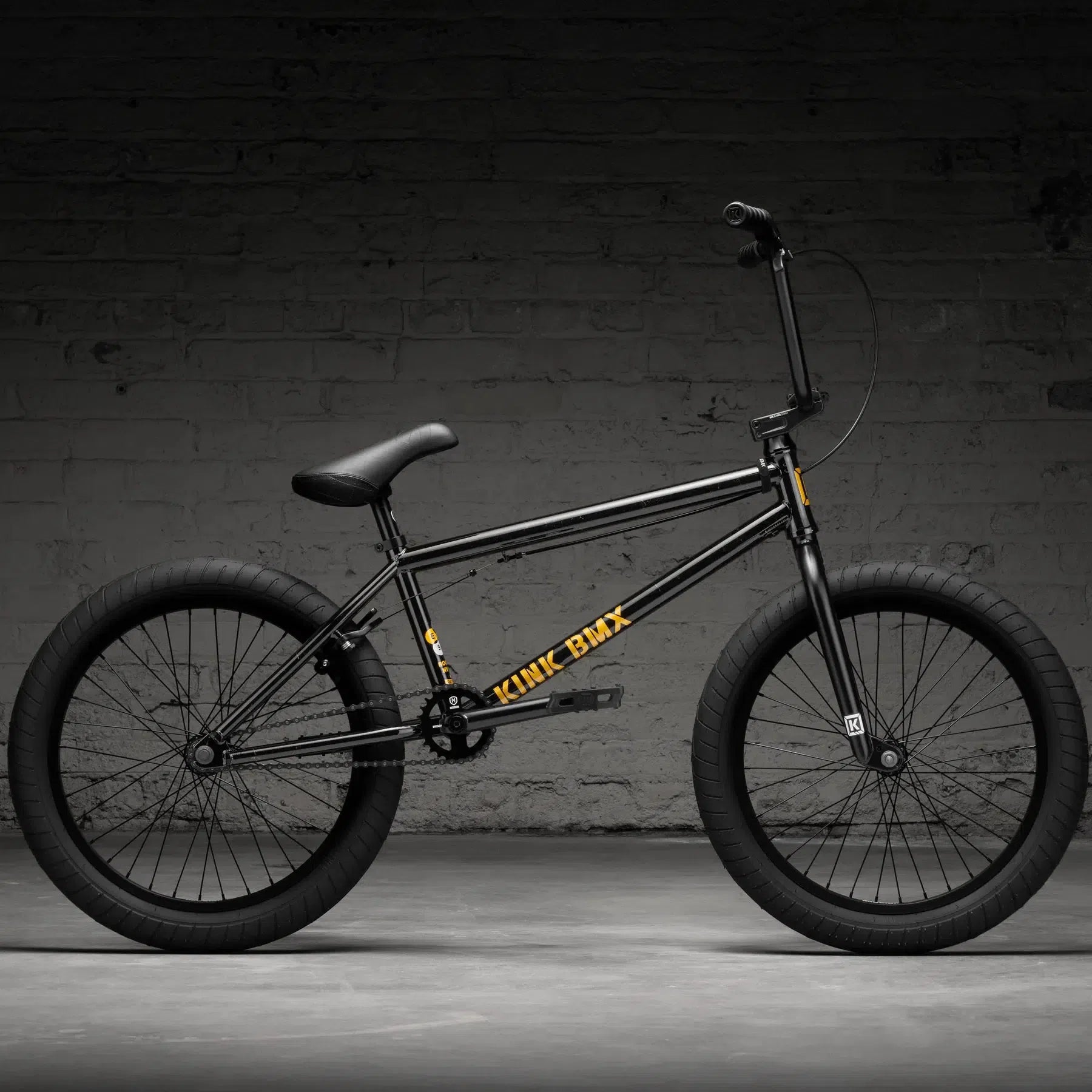 A black Kink Gap XL 20 Inch Bike (2026) with a yellow "Kink BMX" logo shines on its matte Chromoly frame, featuring robust black handlebars and thick tires, set against a dark brick wall backdrop.