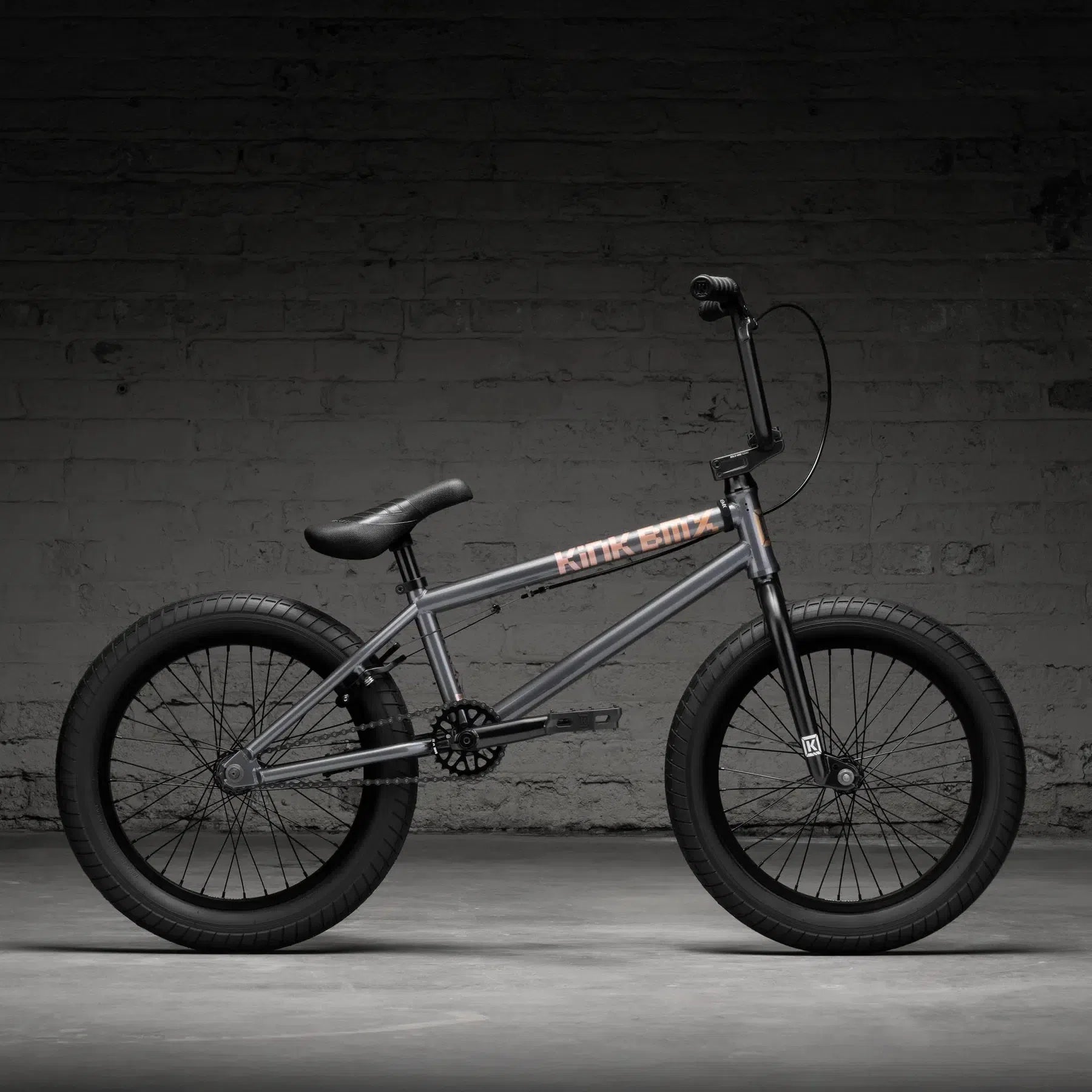 The Kink Kicker 18 Inch Bike (2026) with a gray frame and black tires leans against a brick wall in dim lighting, highlighting its freestyle capabilities.