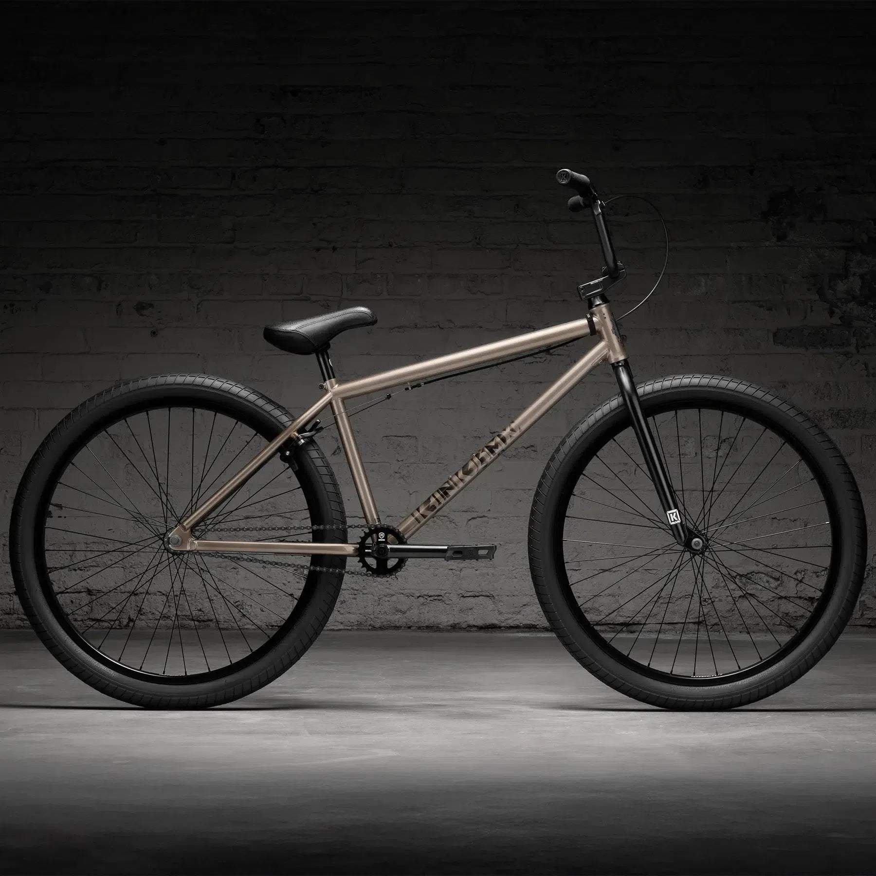 Set against a dimly lit brick wall, the beige Kink Drifter 26 Inch Bike (2026) boasts sleek black tires and handlebars, all built on a robust Chromoly frame.