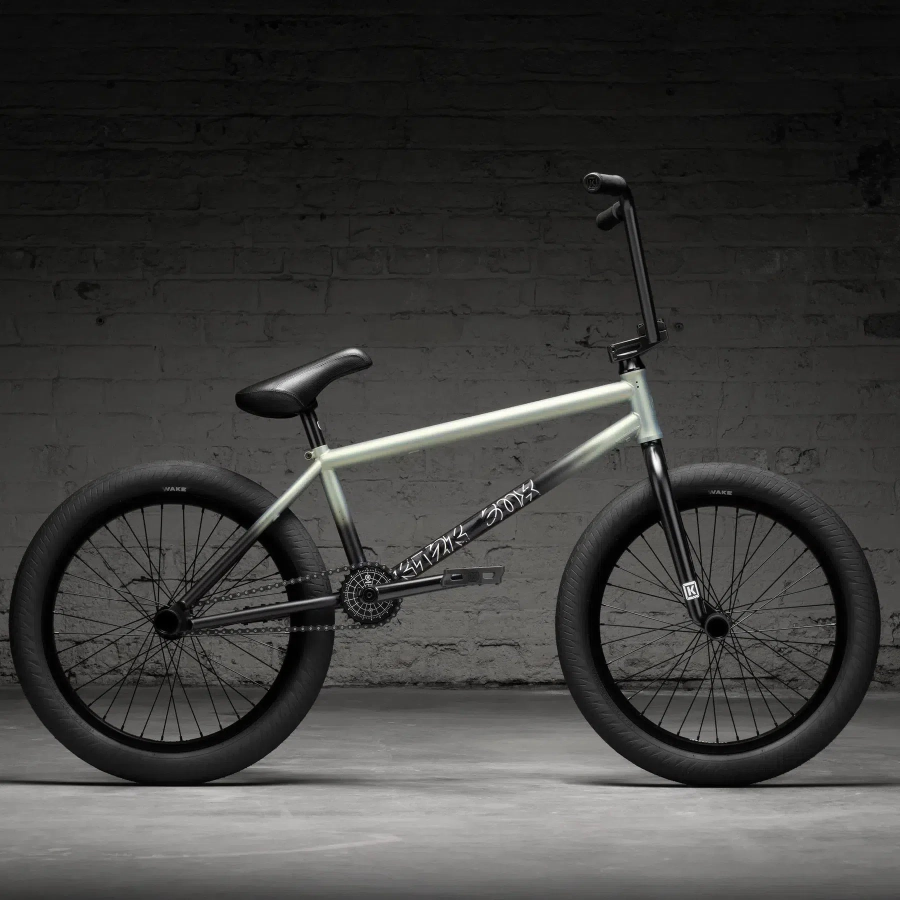 The Kink Switch 20 Inch Bike (2026), featuring a light-colored chromoly frame with black handlebars, seat, and wheels, is showcased against a dark brick wall backdrop.