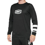 A person wearing the 100% R-Core Youth Jersey in black, which features a long-sleeve design with a "100%" logo and distinctive white stripes on one sleeve, crafted with fade-resistant graphics for lasting durability.