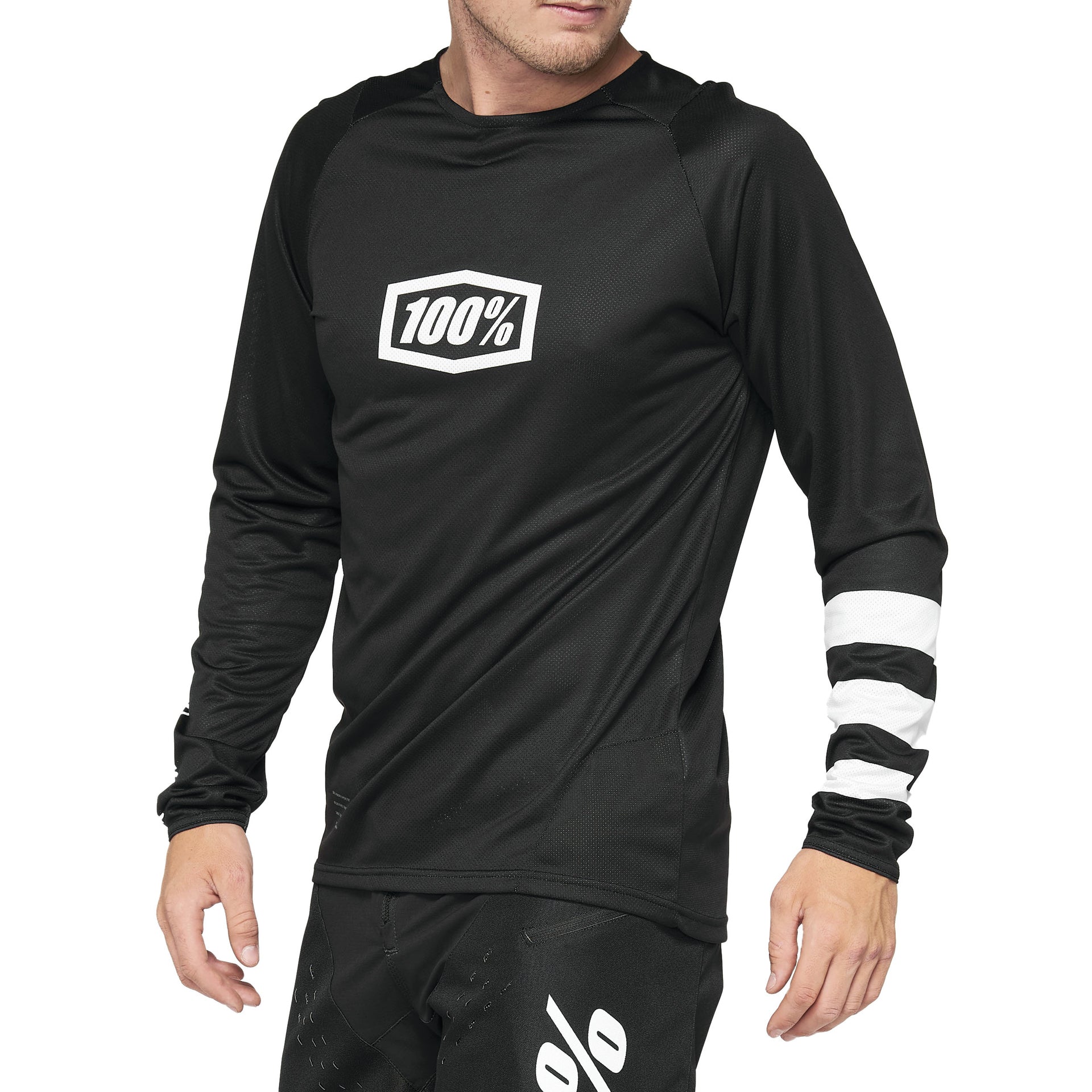 The person is wearing a 100% R-Core Youth Jersey in black, showcasing the "100%" logo on the chest and adorned with a striped sleeve, paired with matching black pants.