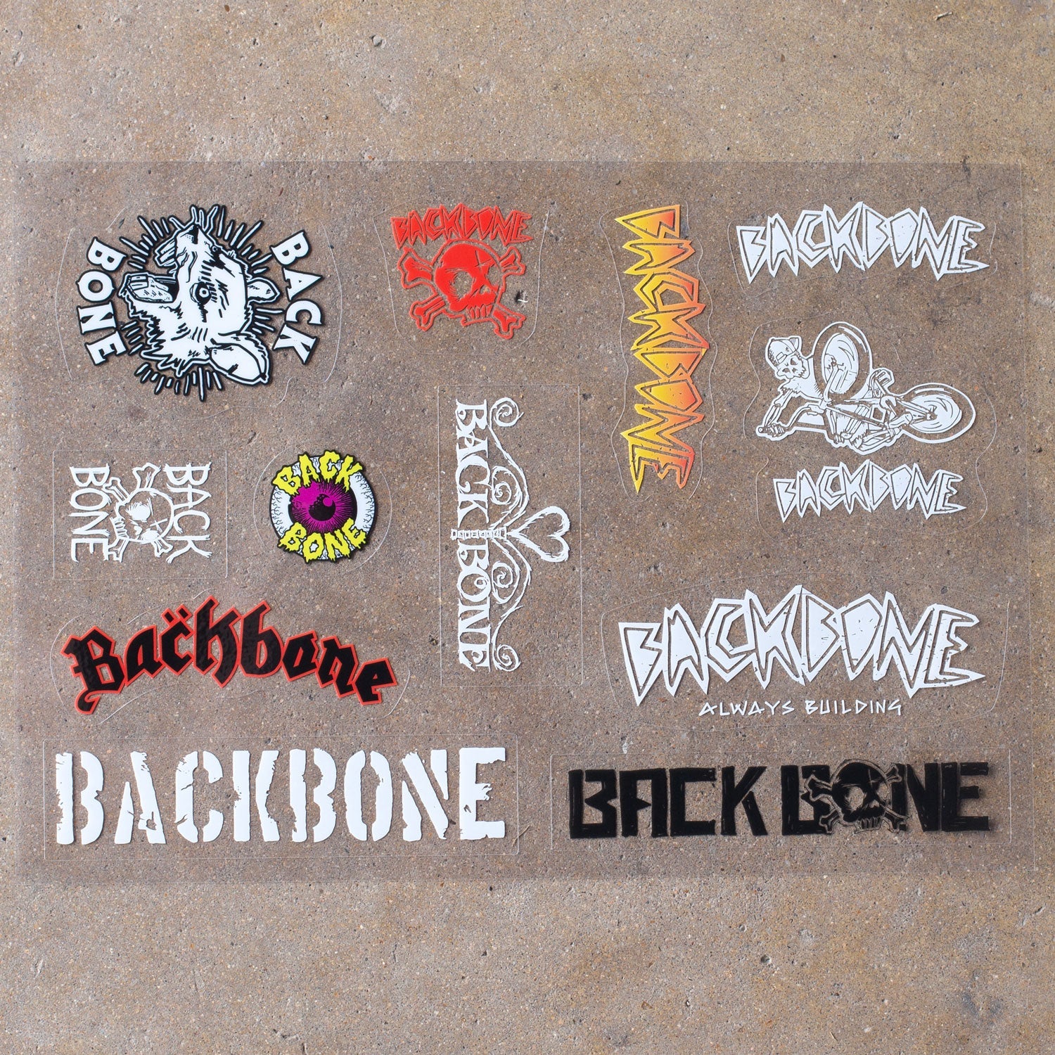 A diverse array of uniquely styled "Backbone Sticker Sheet" BMX stickers in vibrant fonts and colors, artistically arranged on a brown surface.