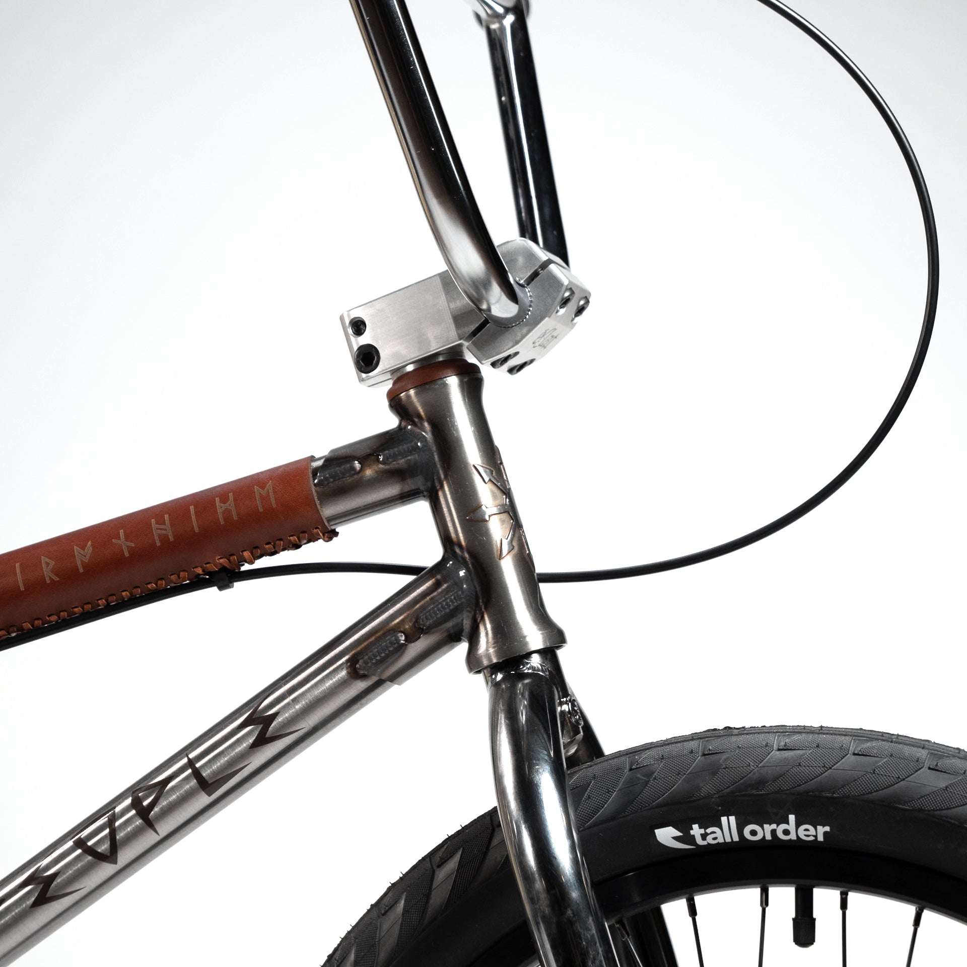 Close-up of the Wethepeople X LUX LAB 'Ironhide' Chaos Machine 20 Inch Custom Bike's front frame and tire. The frame features a distinctive design with Nordic runes and a leather wrap. The Eclat Controller Bars add an extra layer of style, while the tire proudly displays the "tall order" branding.