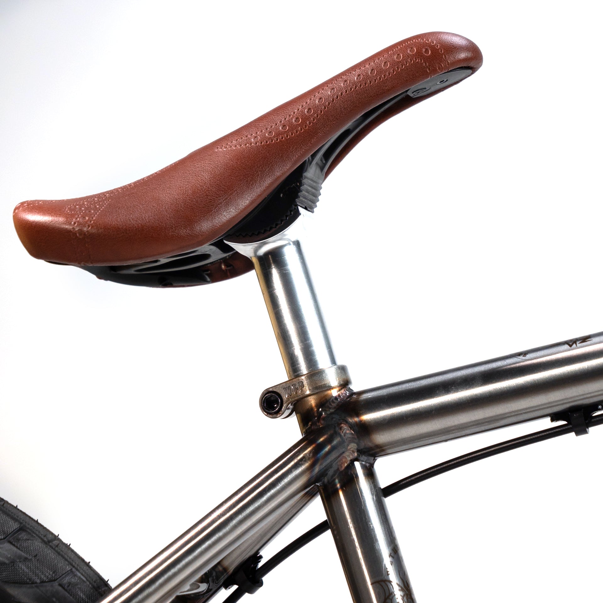 Close-up of the bicycle seat and seat post on a silver frame, featuring a brown leather saddle and a partially visible rear tire—crafted with precision akin to the Wethepeople X LUX LAB 'Ironhide' Chaos Machine 20 Inch Custom Bike.