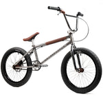 A Wethepeople X LUX LAB 'Ironhide' Chaos Machine 20 Inch Custom Bike, featuring silver frame with brown handlebar grips and seat, black tires, and no visible front or rear brakes, displayed against a white background.