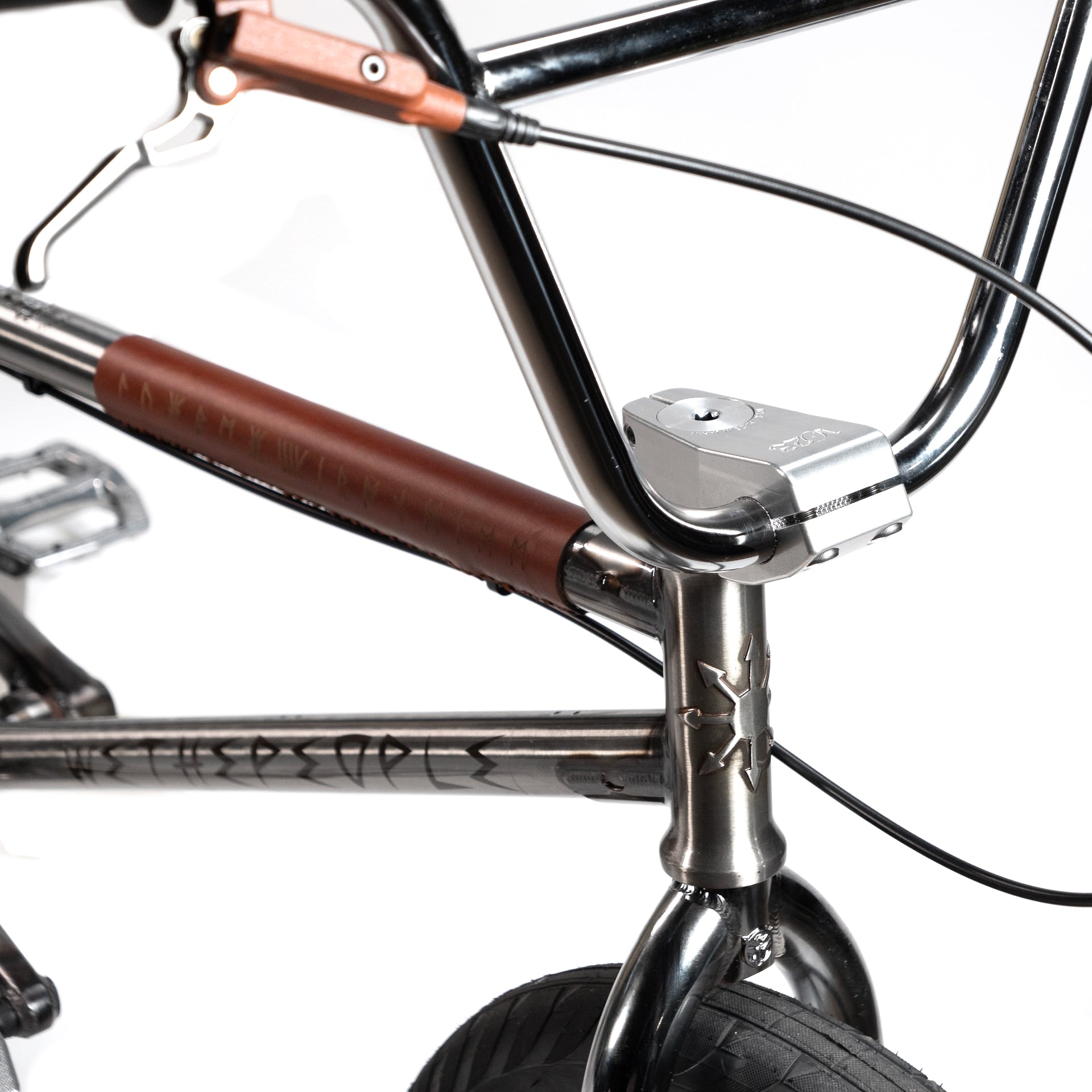 Close-up of the front handlebar area of the Wethepeople X LUX LAB 'Ironhide' Chaos Machine 20 Inch Custom Bike, showcasing Eclat Controller Bars, brake cables, metallic frame, and a partially visible front tire. The frame features a brown leather grip with engraved text.