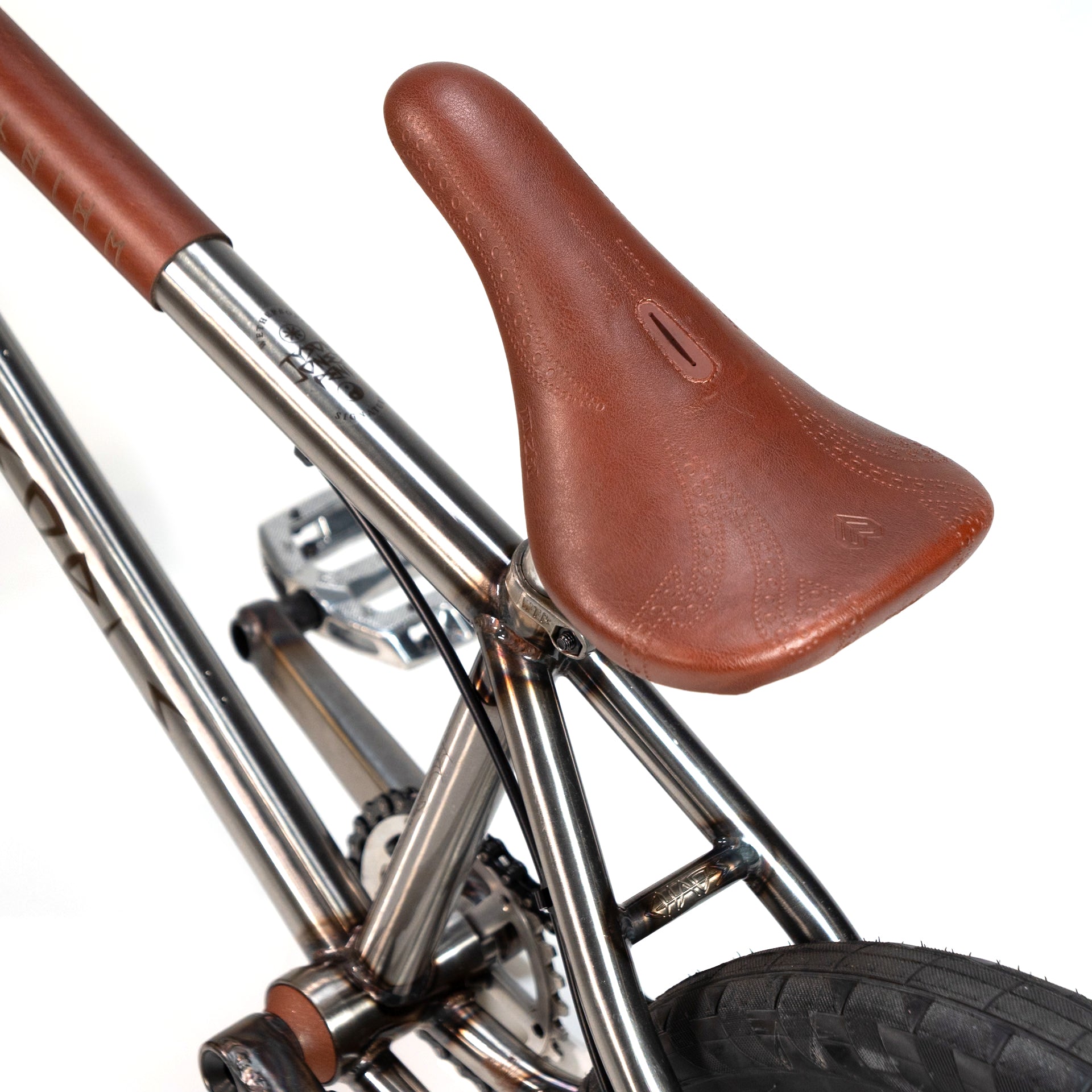 Close-up of a brown leather bike saddle attached to a metal-framed bicycle, featuring the Wethepeople X LUX LAB 'Ironhide' Chaos Machine 20 Inch Custom Bike rear wheel and pedal assembly.