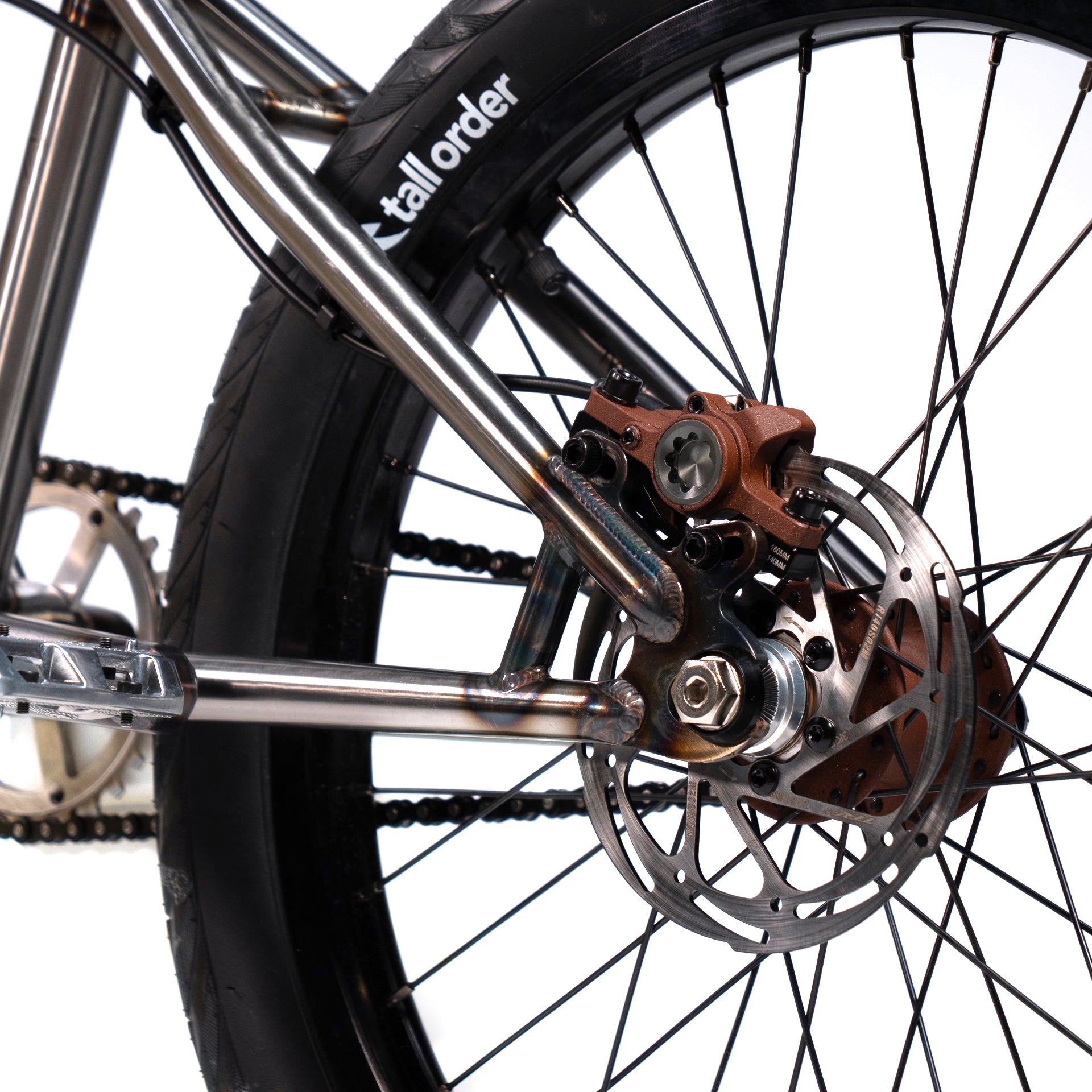 Close-up of a bicycle rear wheel, highlighting the disc brake rotor and caliper. The bike frame, equipped with Wethepeople X LUX LAB 'Ironhide' Chaos Machine 20 Inch Custom Bike elements, and tire are partially visible.