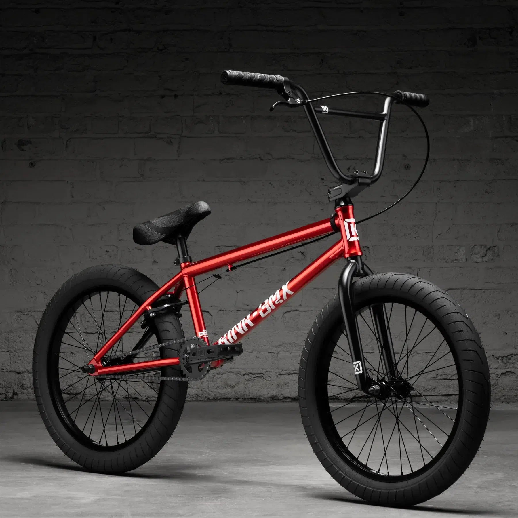 A red Kink Launch 20 Inch Bike (2026) with Mission parts stands against a textured gray brick wall, boasting black handlebars and tires for a sleek look, enhanced by durable sealed bearings.