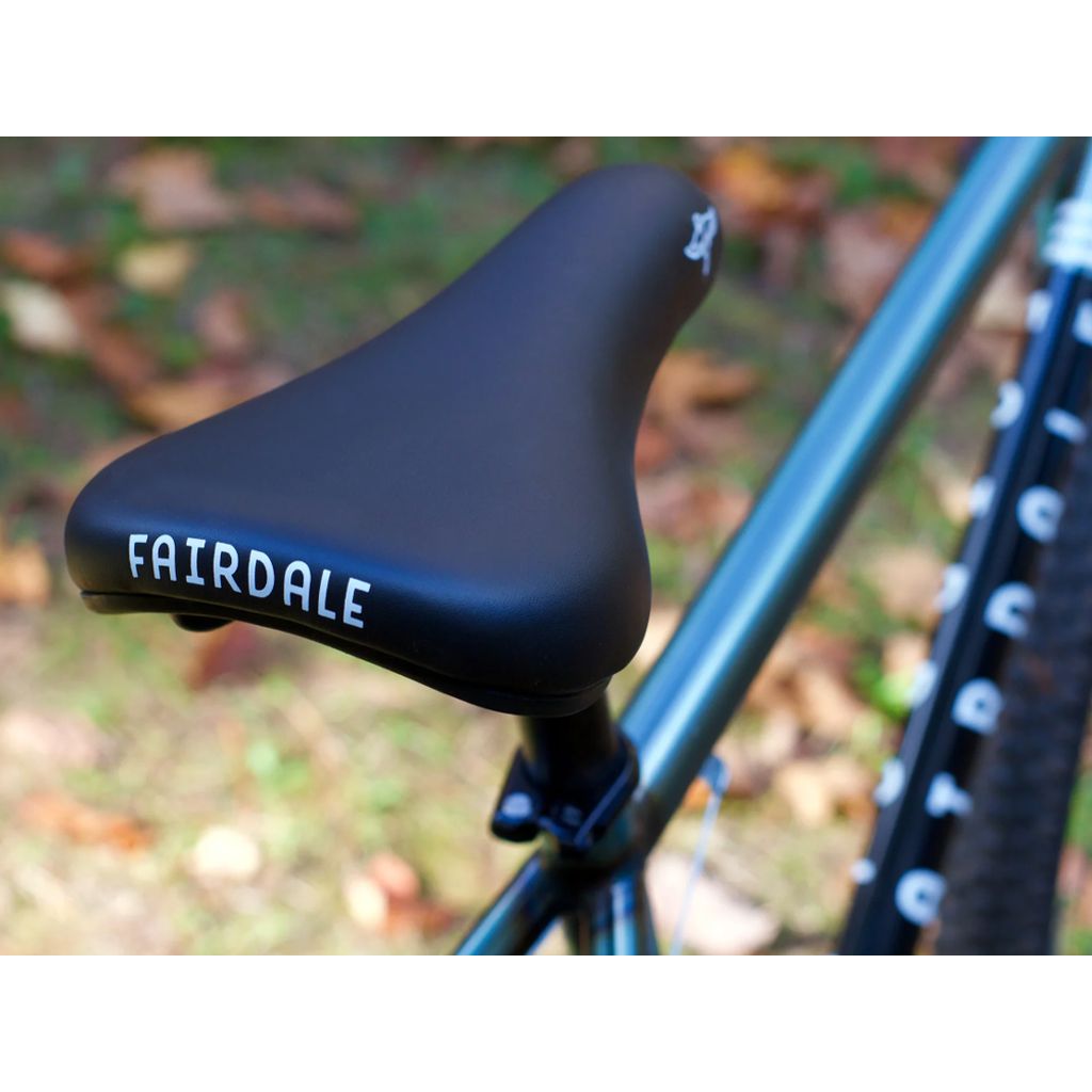 A close-up of the Fairdale Taj 27.5 Inch Bike (2022) seat with the brand "Fairdale" prominently displayed, accentuating the bike's sleek design. The partially visible frame and rear wheel highlight its sturdy chromoly construction against a backdrop of fallen leaves.