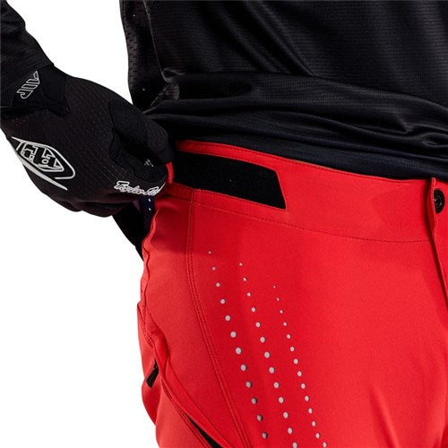 A person adjusts the waistband of the TLD 25.1 Sprint Pant / Mono Fiery Red with a gloved left hand, highlighting its durable materials and race-ready design.