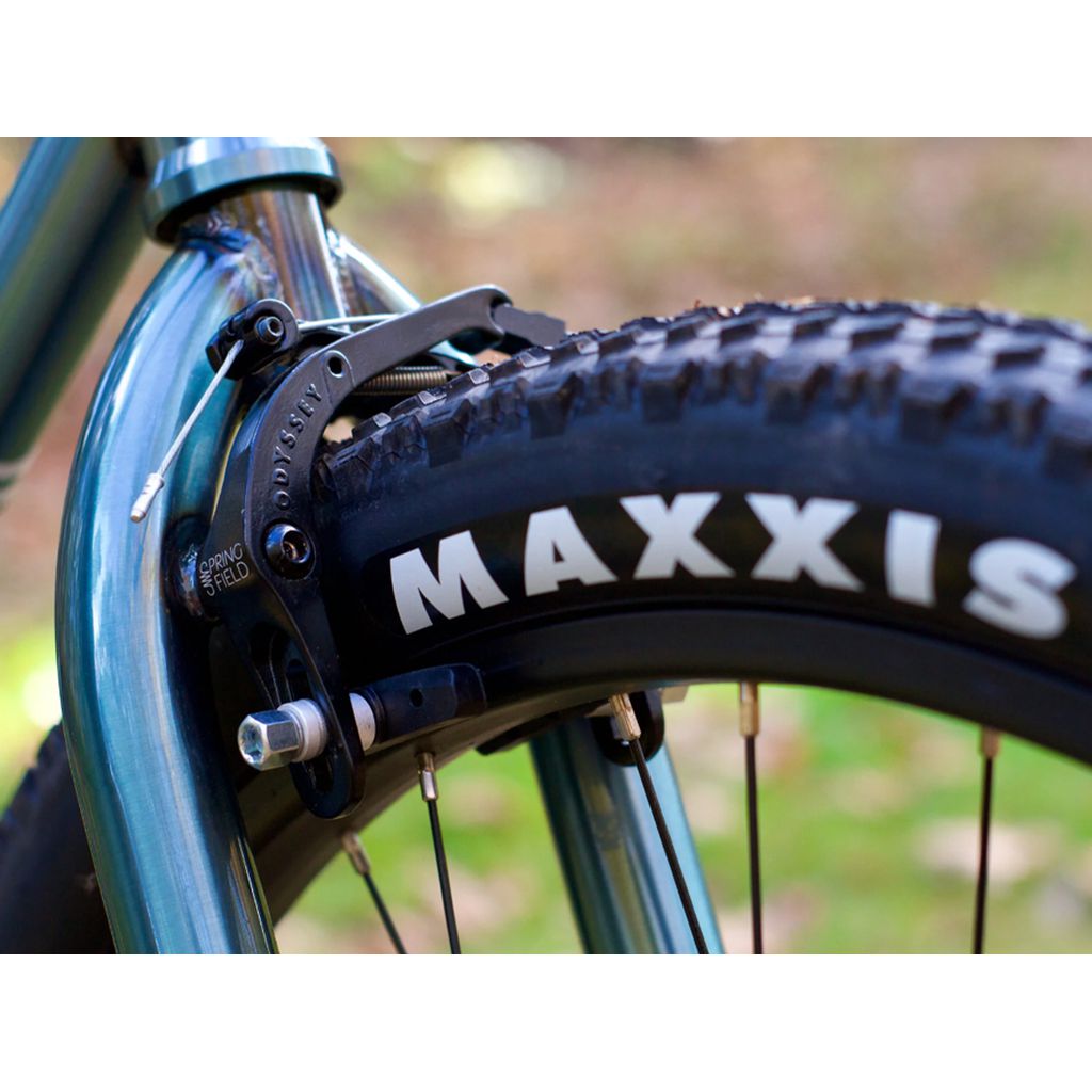 Close-up of a Fairdale Taj 27.5 Inch Bike (2022)'s front wheel featuring a Maxxis tire. The visible blue chromoly frame and black brake caliper highlight its sleek design and single-speed drivetrain.