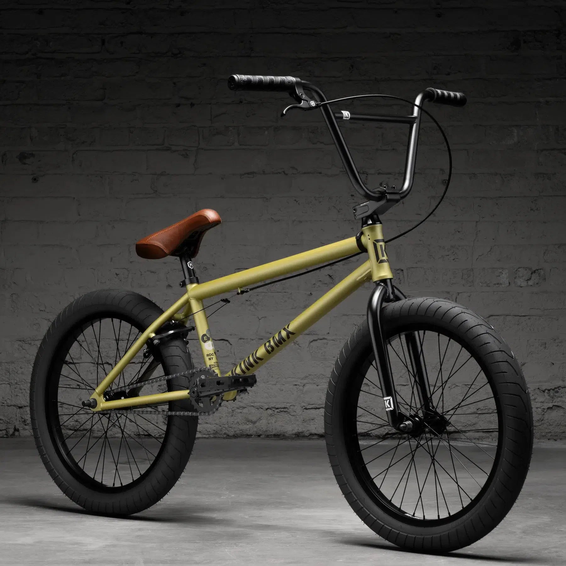 The Kink Gap XL 20 Inch Bike (2026) with a yellow frame, black handlebars and wheels, and a brown seat is displayed against a textured gray brick wall. Its Chromoly build is ideal for urban adventures.