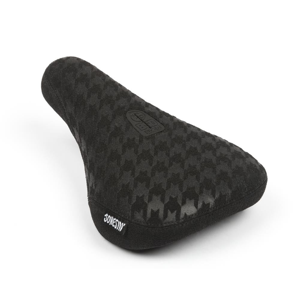 The BSD Jonesin Pivotal Seat is a black fat bicycle seat with a houndstooth pattern and Sam Jones signature, featuring Kevlar bumpers for durability. The brand logo is elegantly on top, and the seat is angled to showcase its stylish side and back.