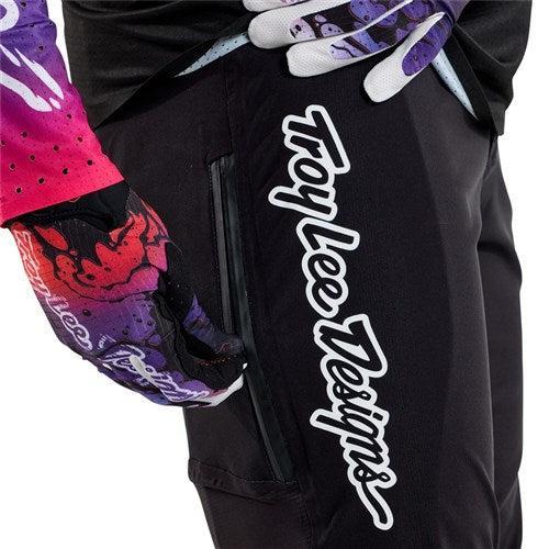 Close-up of a person wearing TLD 25.1 Sprint Ultra Pant / Mono Black with "Troy Lee Designs" logo and colorful gloves, highlighting the precision fit as they unzip a pocket made from Bluesign-approved fabric.