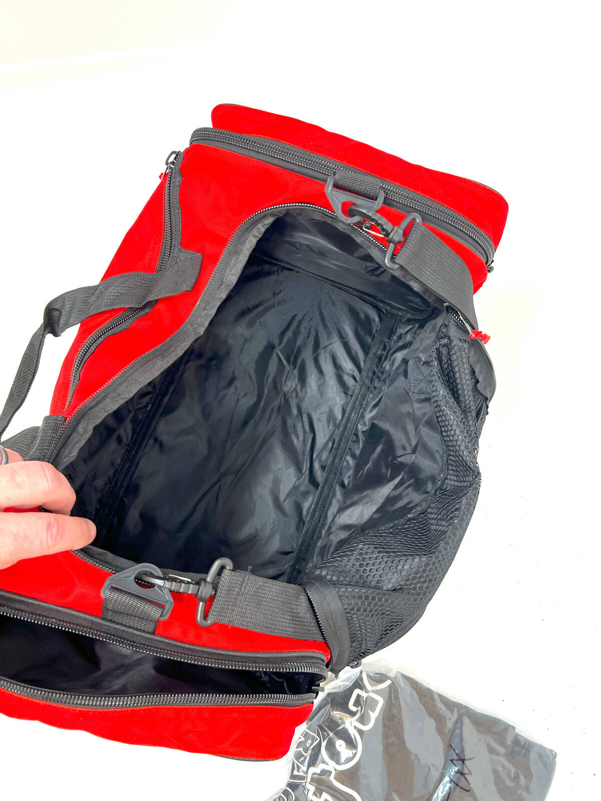 A Profile Racing Custom Team Gear Bag, made from durable 600D polyester, sits with open compartments. A hand reaches to open the bag, while nearby a pack of small plastic bags can be seen.