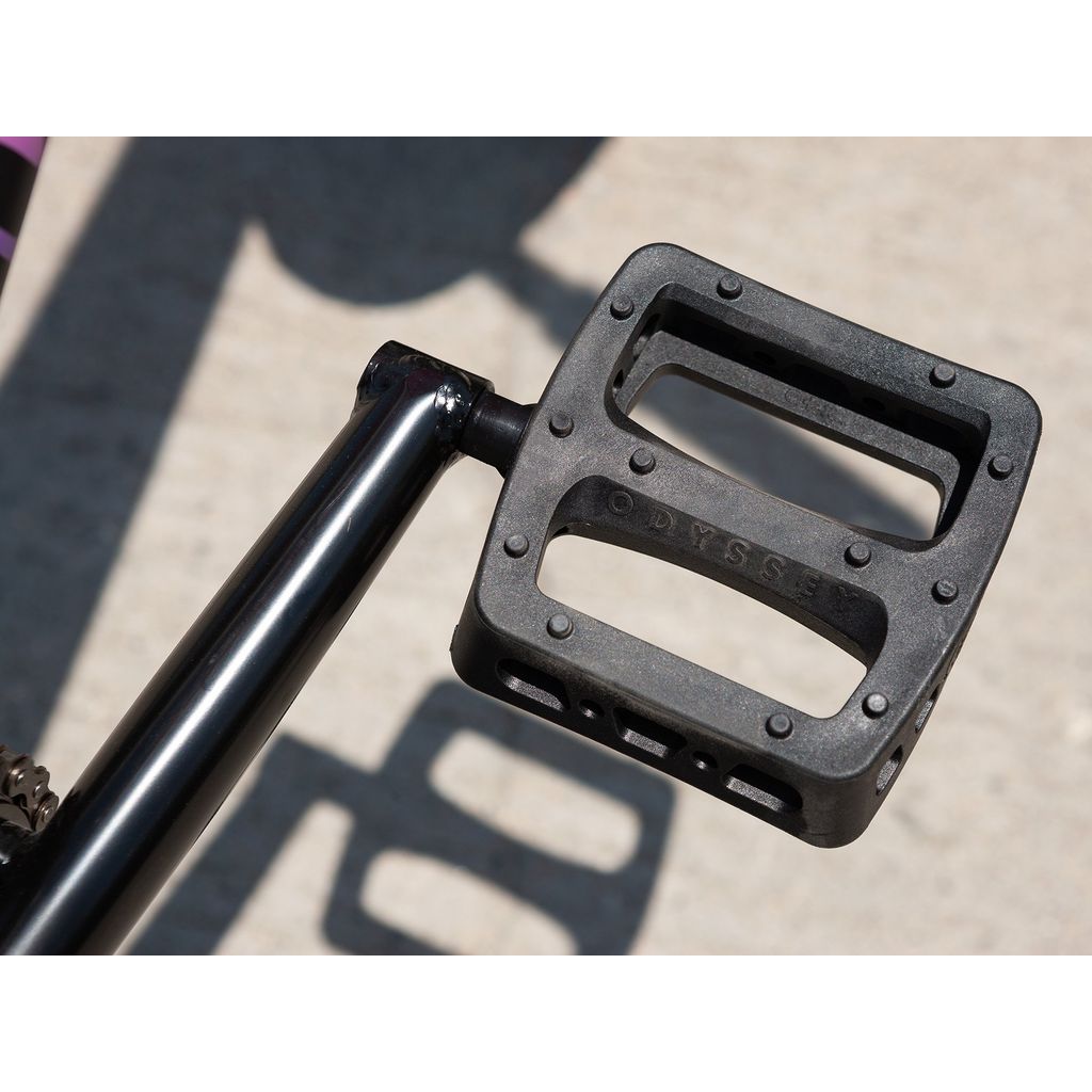 Close-up of a black BMX bike pedal featuring "Odyssey" branding, attached to a crank arm, resting on a concrete surface and ideal for the Sunday Street Sweeper 20 Inch Bike.