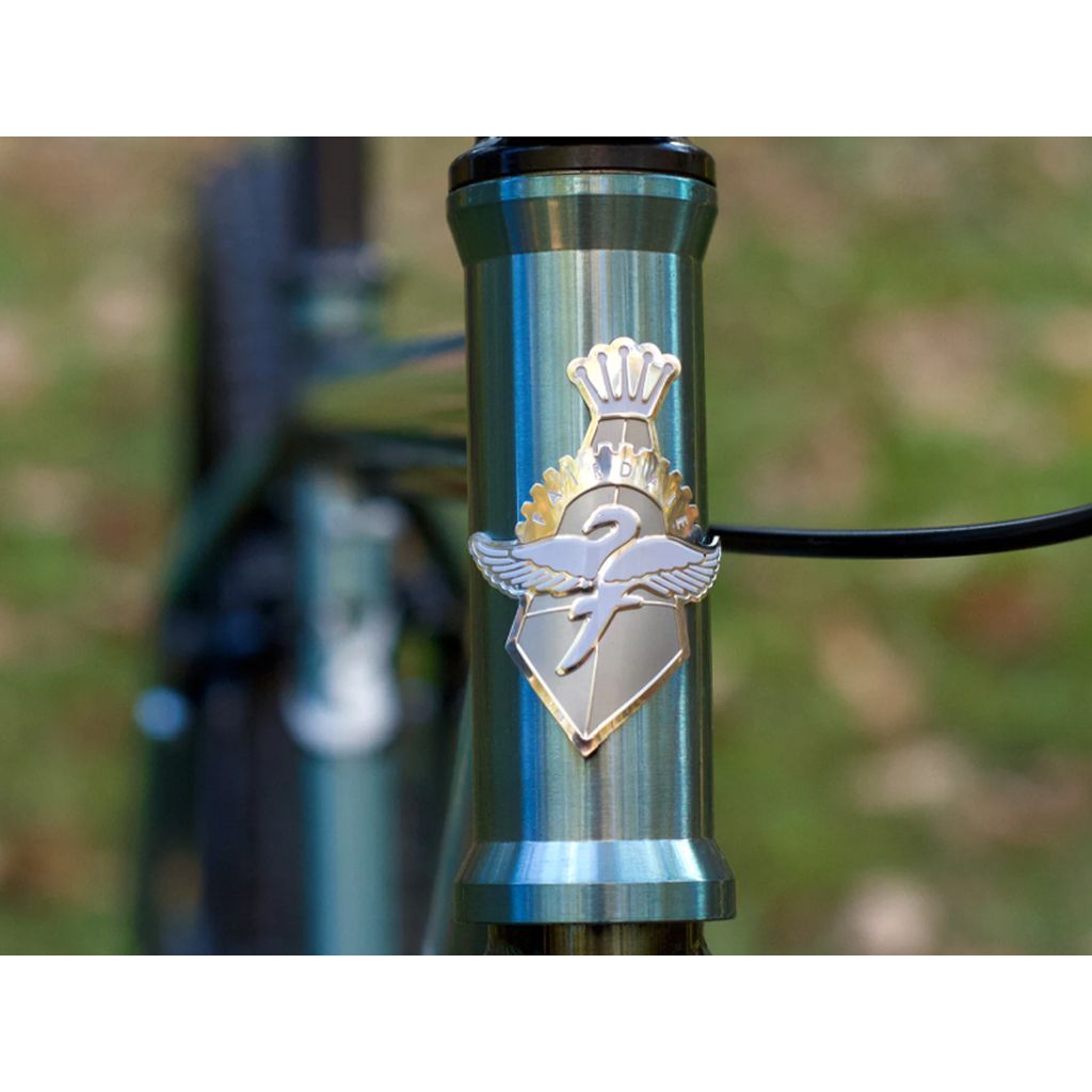 A close-up of the metallic head tube from the Fairdale Taj 27.5 Inch Bike (2022) showcases a crowned winged emblem in silver and gold. The chromoly frame is flawlessly complemented by its single-speed drivetrain, with blurred greenery in the background adding a natural element to this sleek design.