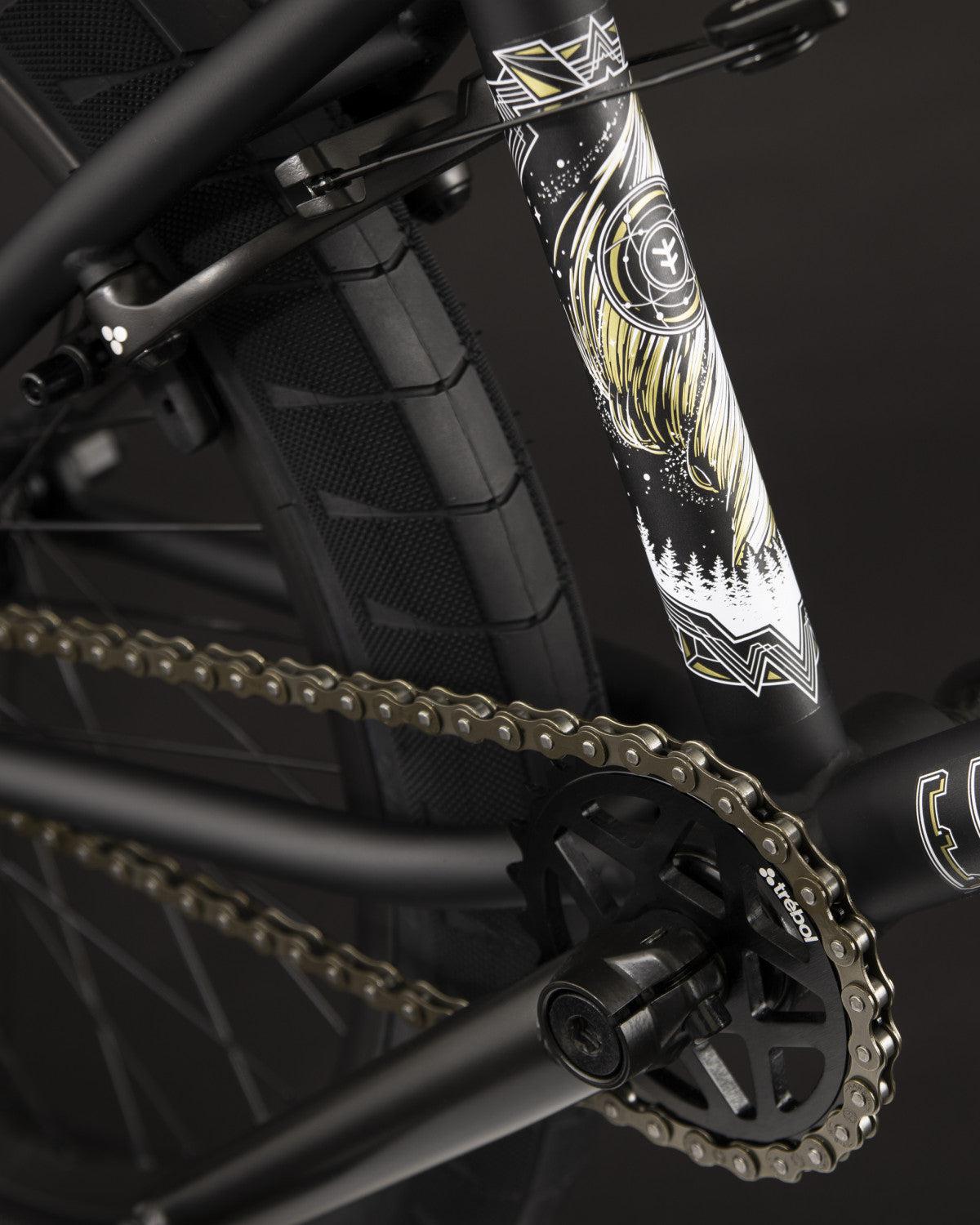 A close-up of the Fly Bikes Electron 20 Inch Bike highlighting its decorated frame made from Cr-Mo material, equipped with Trebol Tires, a shimmering gold chain, and intricate graphics featuring a nature-themed design.