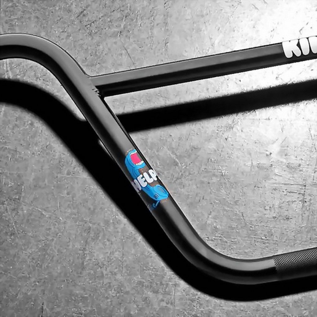 A Kink Williams Bars BMX handlebar with a blue sticker and the "Kink" logo rests on a textured gray surface, showcasing its sturdy chromoly tubing enhanced by TruTherm heat treatment.