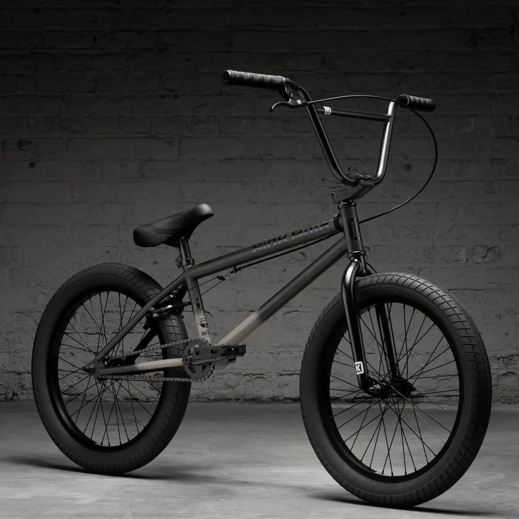 The Kink Curb 20 Inch Bike (2026), featuring a sturdy matte-black frame, wide tires, and high handlebars, leans against a textured gray brick wall—ideal for entry-level riders.