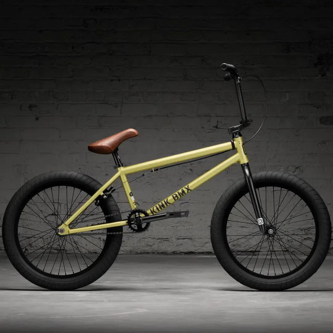 A yellow Kink Gap XL 20" BMX bike with thick black tires leans against a brick wall, featuring a brown seat and sleek black handlebars.