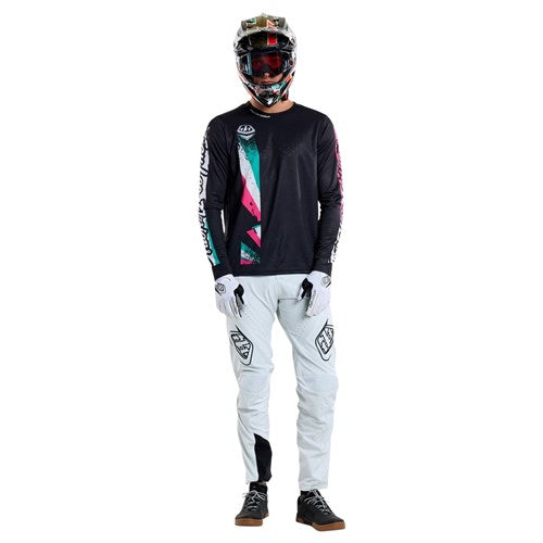 A person in a motocross outfit stands confidently against a plain backdrop, wearing a black helmet, TLD 25.1 Sprint Jersey / Full Pull Black with colorful accents, white pants adorned with logos, white gloves, and black shoes.