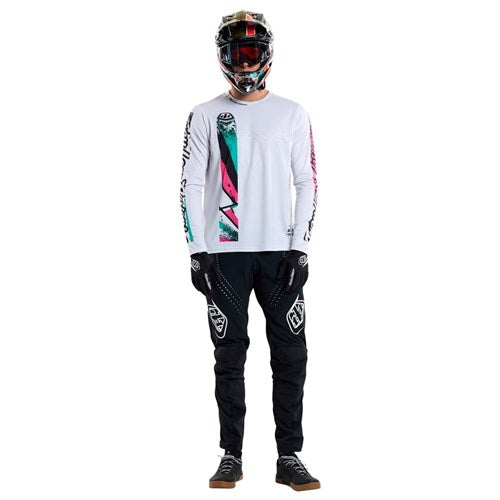 A person confidently poses against a white background, wearing a vibrant motocross outfit featuring a TLD 25.1 Sprint Jersey / Full Pull Chalk, helmet, white long-sleeve top, black pants, gloves, and boots—ready for intense race weekends.