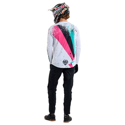 A person in colorful motocross gear stands facing away, wearing a Troy Lee Designs helmet and gloves. The TLD 25.1 Sprint Jersey / Full Pull Chalk features pink, teal, and black accents, paired perfectly with sleek black pants for thrilling race weekends.