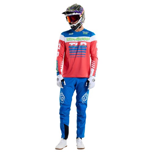 A rider showcases a vibrant motocross ensemble, highlighted by a Troy Lee Designs TLD 25.1 Sprint Jersey in Fifty-50 Fiery Red, paired with blue pants and gloves, set against a white backdrop—ideal for exciting race weekends.