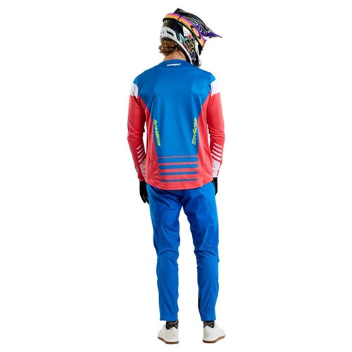 With their back to the camera, a person wears a vibrant TLD 25.1 Sprint Jersey in Fifty-50 Fiery Red from Troy Lee Designs, holding a helmet—an iconic look for thrilling race weekends.