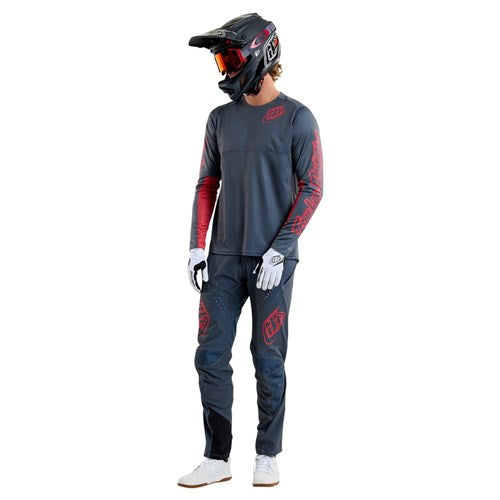 A person in a dark Troy Lee Designs Sprint Jersey with red accents, helmet, goggles, gloves, and sneakers stands against a white background.