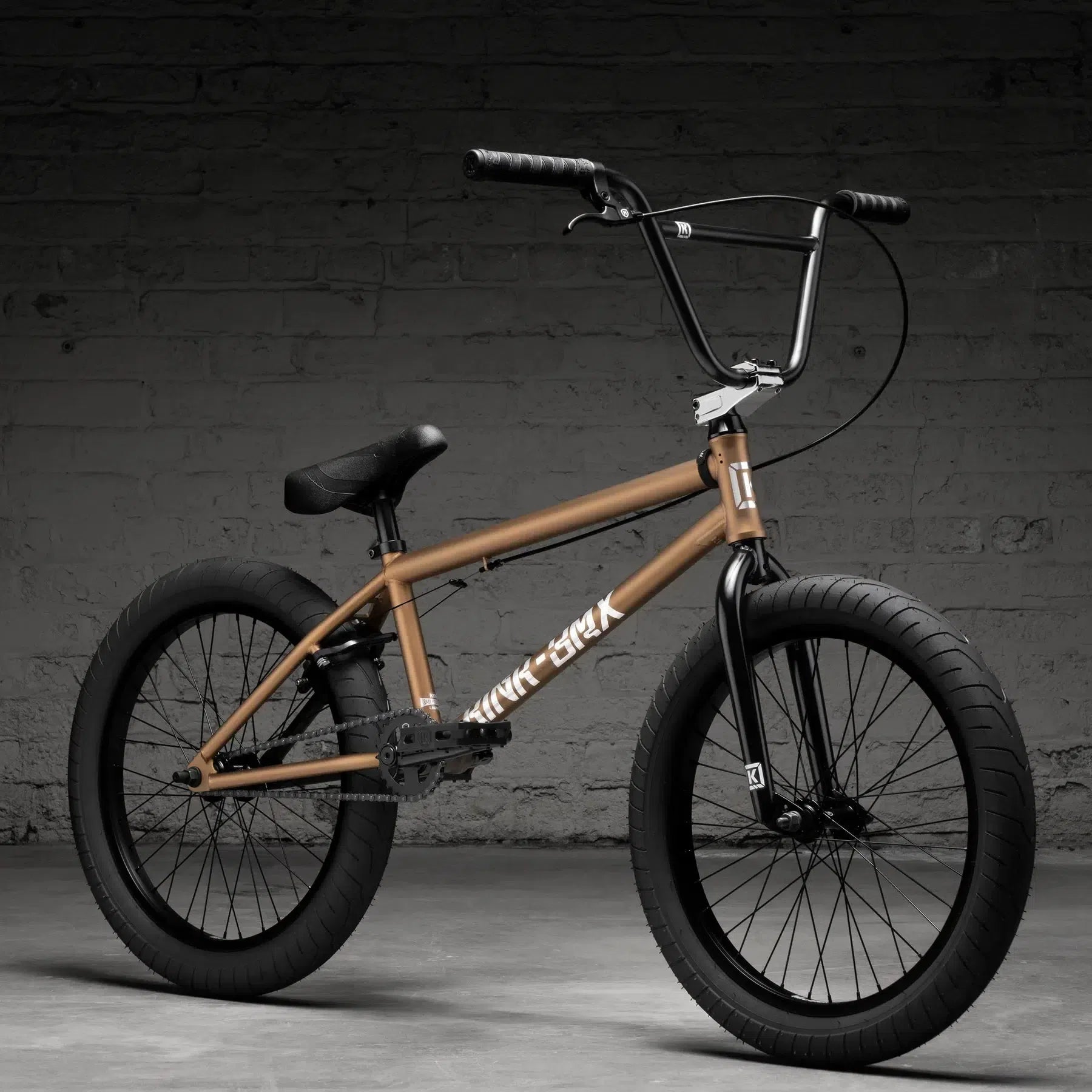 A Kink Launch 20 Inch Bike (2026), featuring thick black tires, Mission parts, and smooth-rolling sealed bearings, is positioned on a concrete floor against a textured gray brick wall.