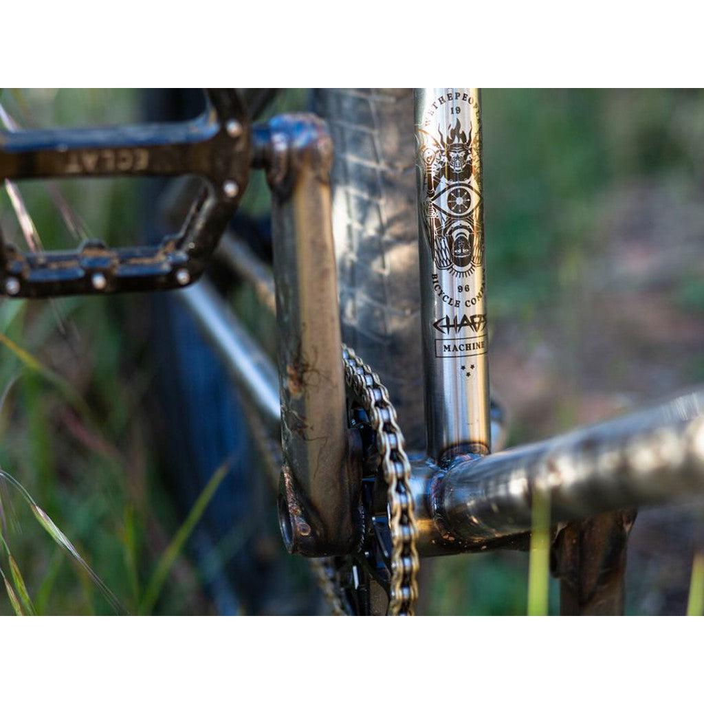 Close-up of a bicycle's pedal, chain, and the Wethepeople Chaos Machine 22 Frame (Tyson Jones-Peni Signature). The bike rests on grass.