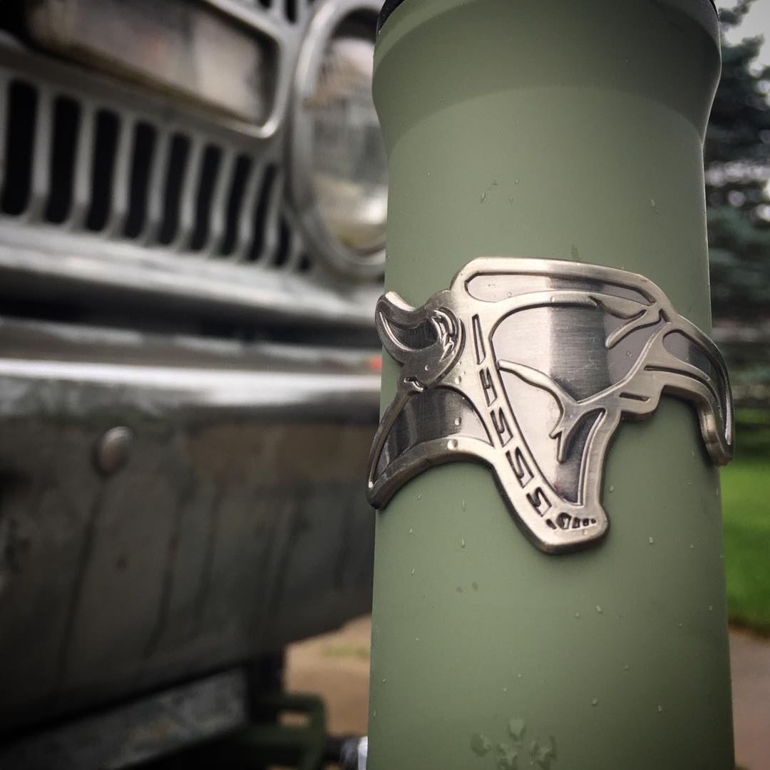 Close-up of a green water bottle with a metal bullhead logo, showcasing its Animal Head Tube Badge design. A vintage vehicle front grille is visible in the blurred background.